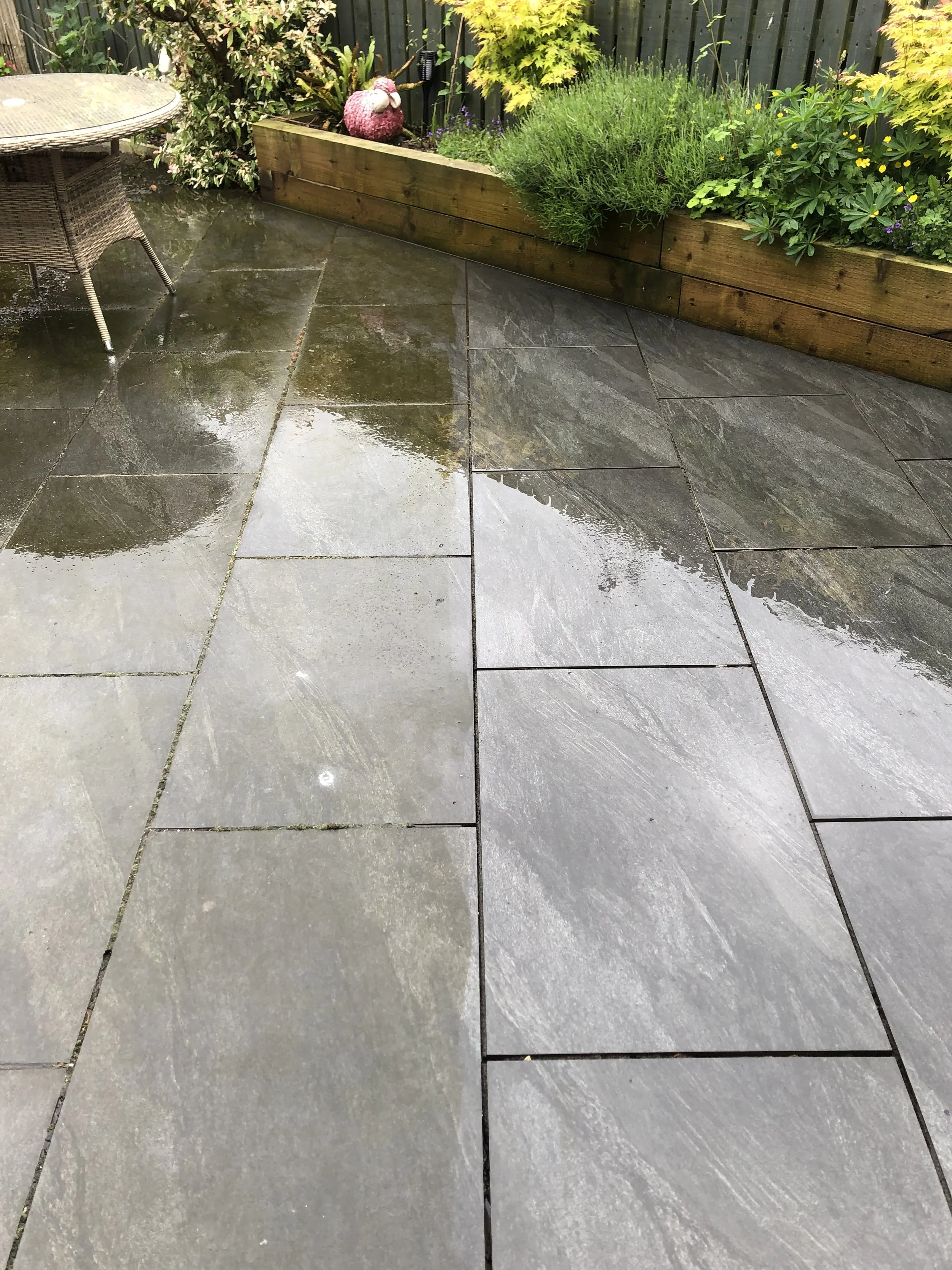 Wet patio with large gray stone tiles, reflected rain, and a background of green garden plants and a wooden border. There is a wicker chair on the left.