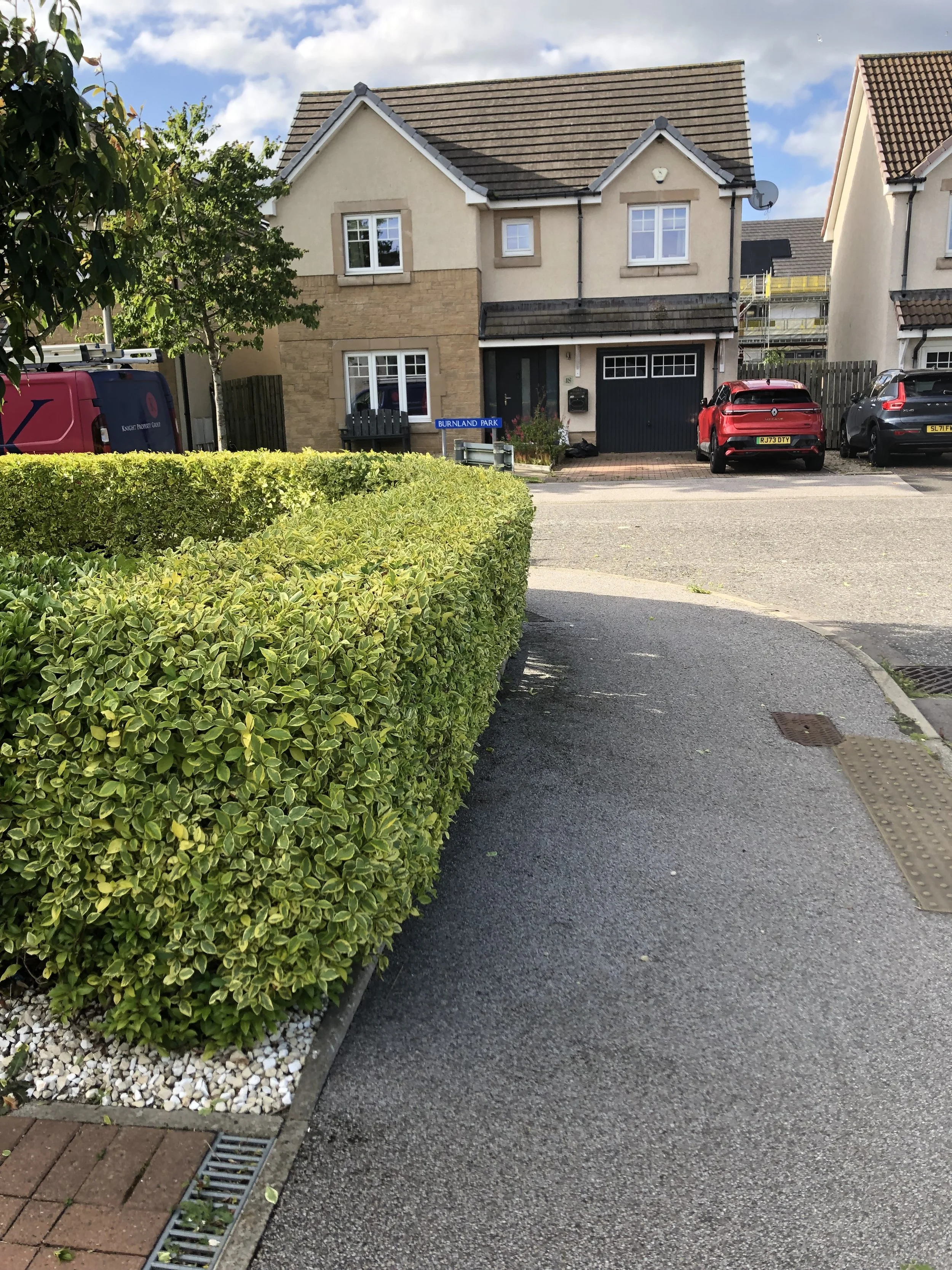 Residential street with well-maintained bushes and houses in the background, showing a driveway, parked cars, and a street sign that reads 'BURNLAND PARK'.