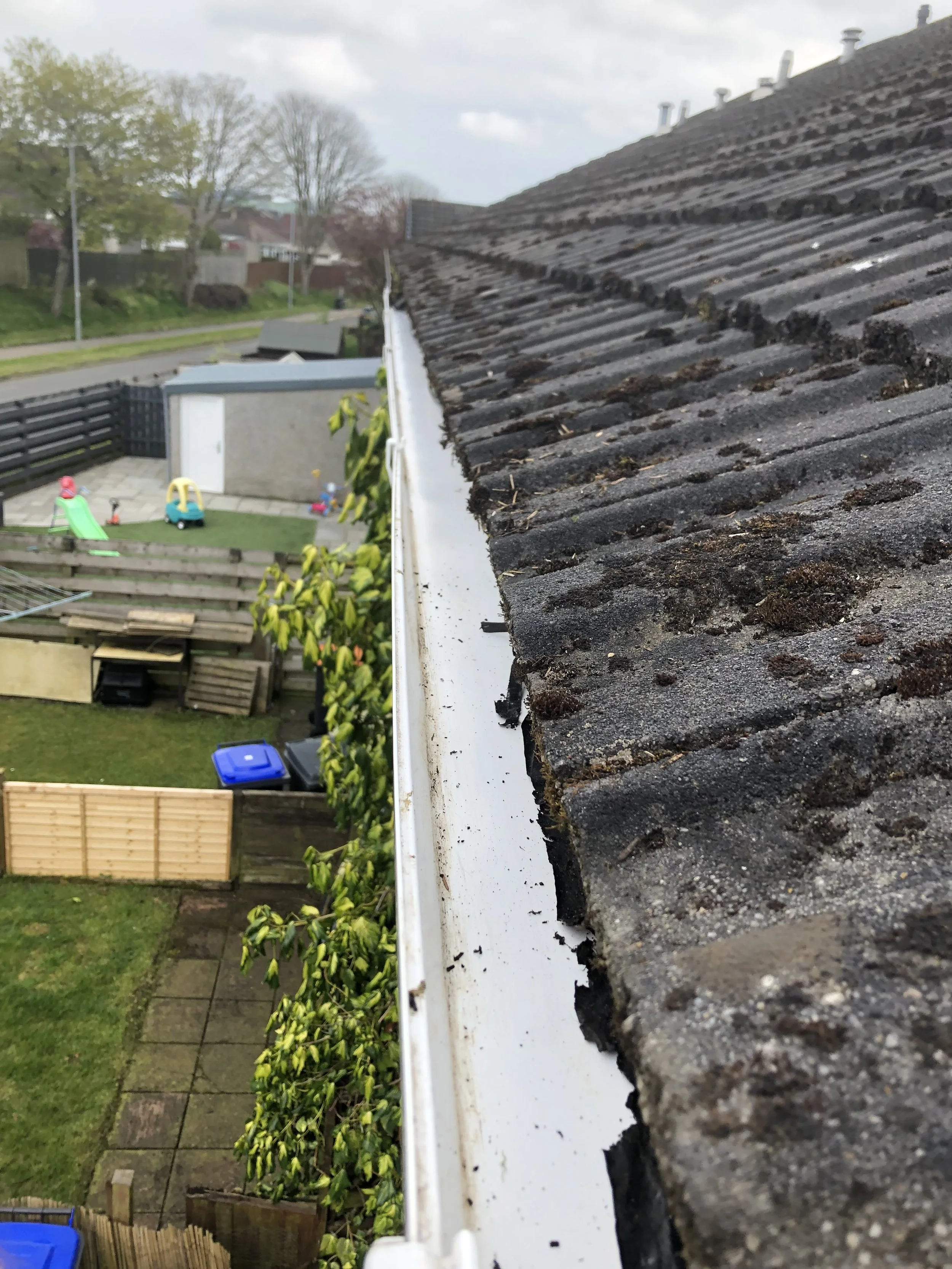 Close-up of a residential house roof with black and gray weathered shingles and a white gutter, overlooking a backyard with a sliding slide, playhouse, and garden.