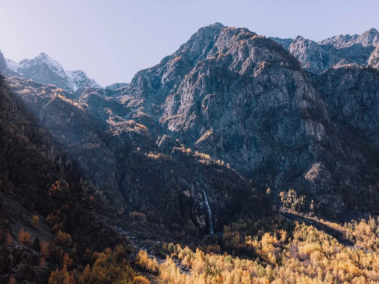 A mountain landscape with rocky peaks, a small waterfall, and autumn-colored trees at the base.