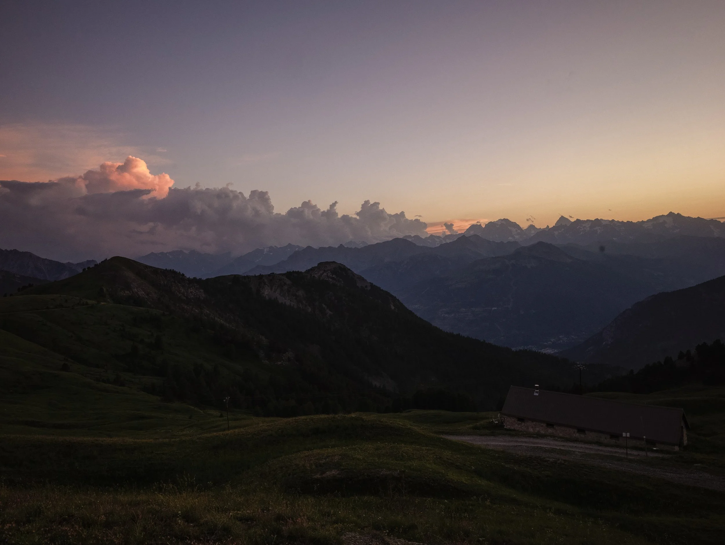A mountain landscape at sunset with dark silhouettes of rolling hills, a barn, and snow-capped mountains in the distance under a colorful sky with clouds.