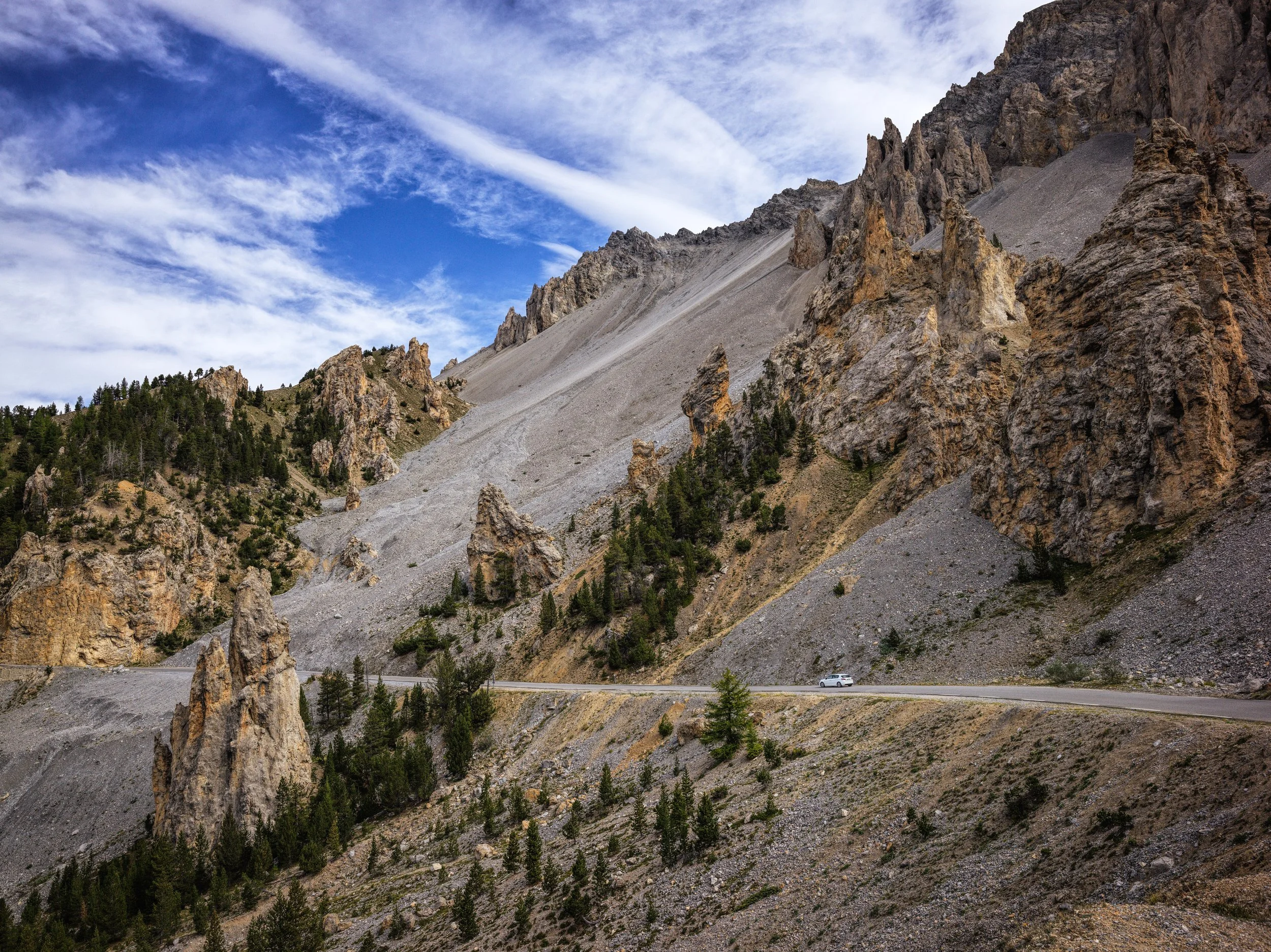 A white vehicle travels on a winding mountain road with rocky cliffs and sparse green trees against a blue sky with scattered clouds.