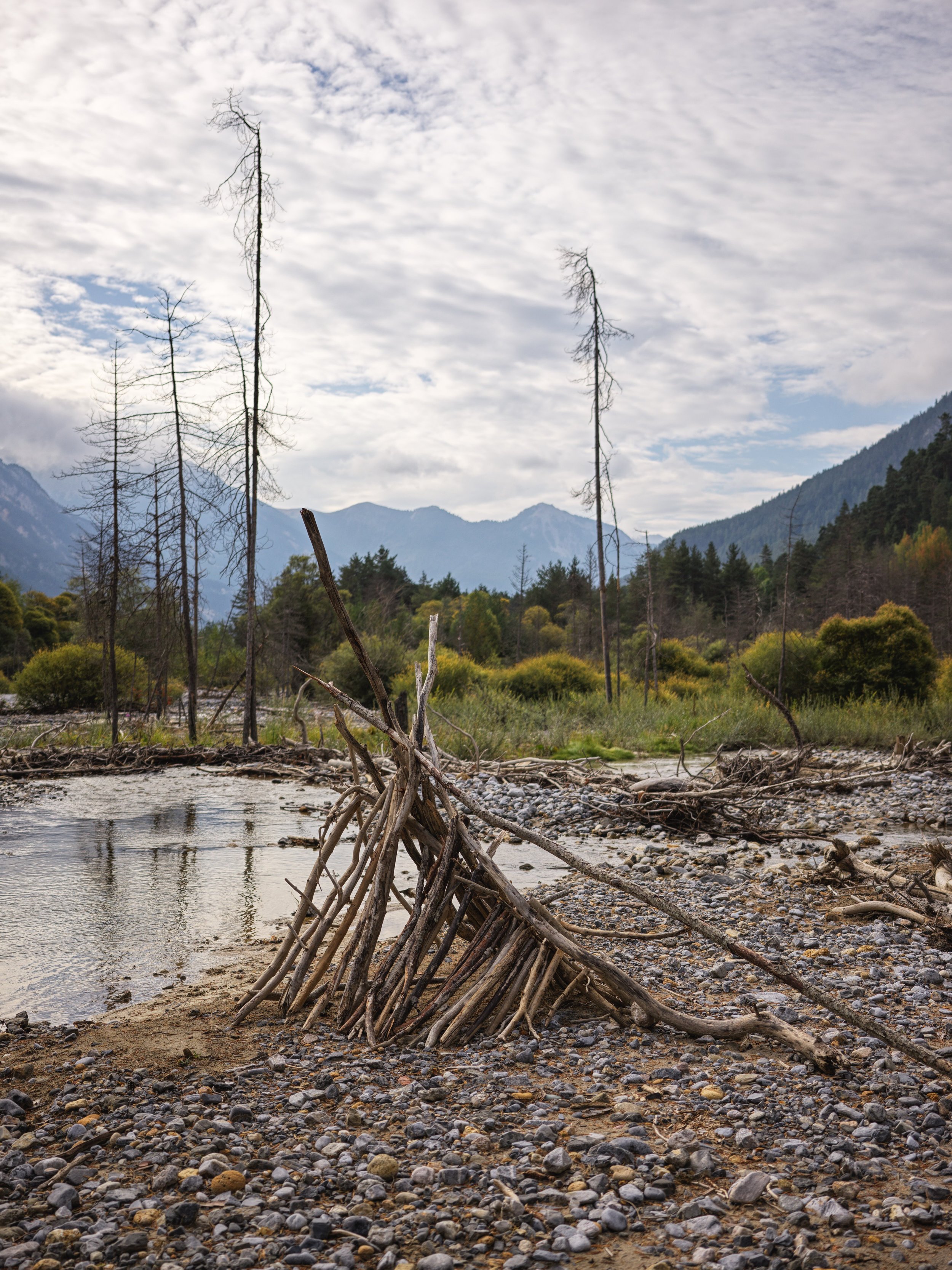 A riverbank with a makeshift teepee structure made of driftwood branches, surrounded by pebbles and trees, with mountains and a cloudy sky in the background.