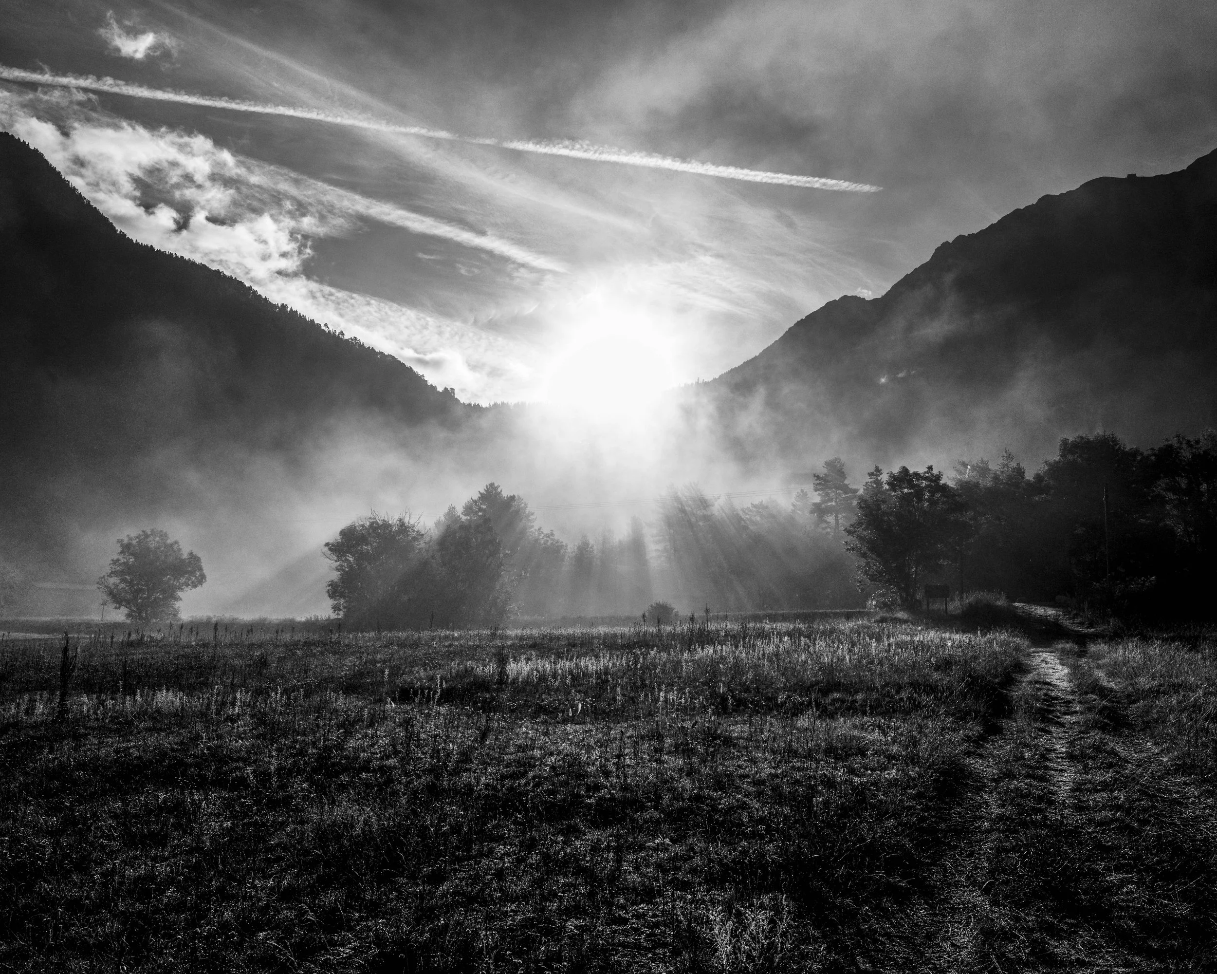 Black and white photograph of a sun rising over a mountain valley with trees, mist, and a dirt path.