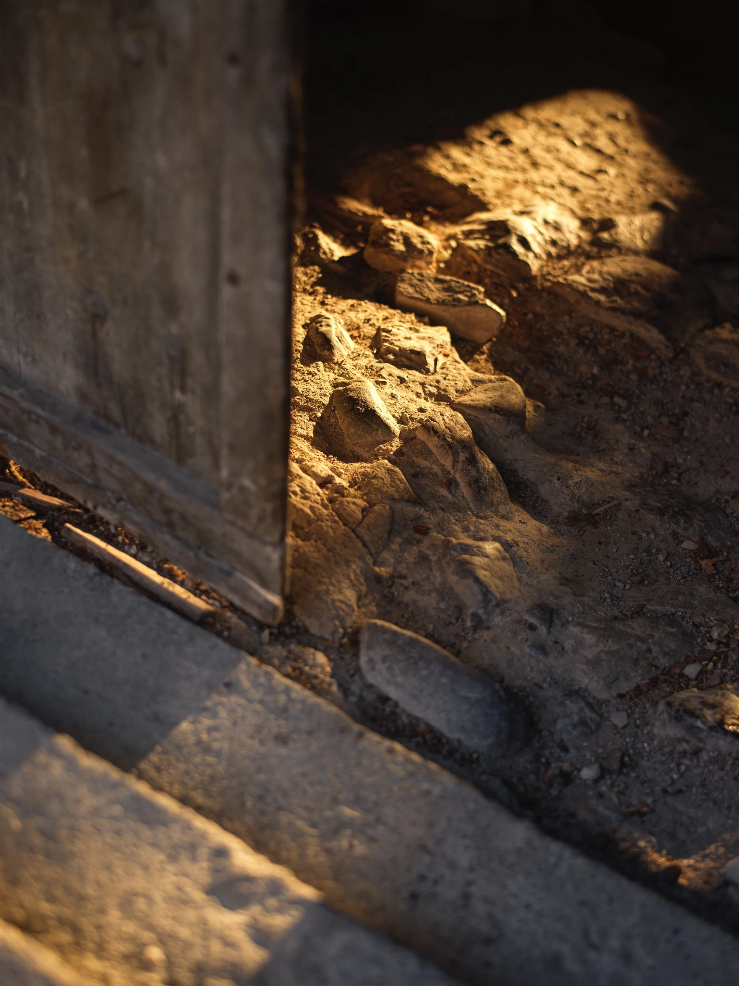 Close-up of a crack with uneven stones and dirt, illuminated by warm light, near a wooden structure.