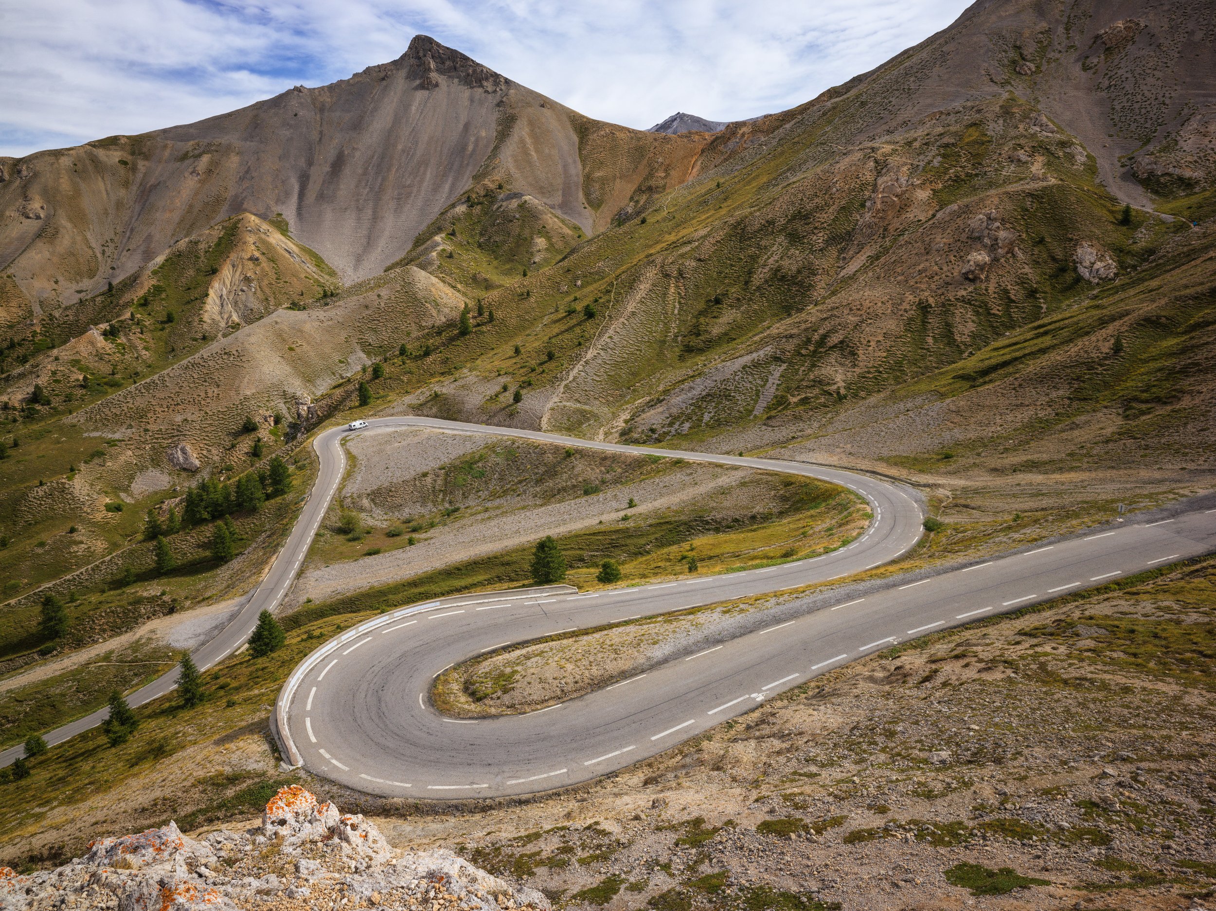 A winding mountain road through rugged, dry terrain with sparse trees and steep slopes, with mountains in the background.