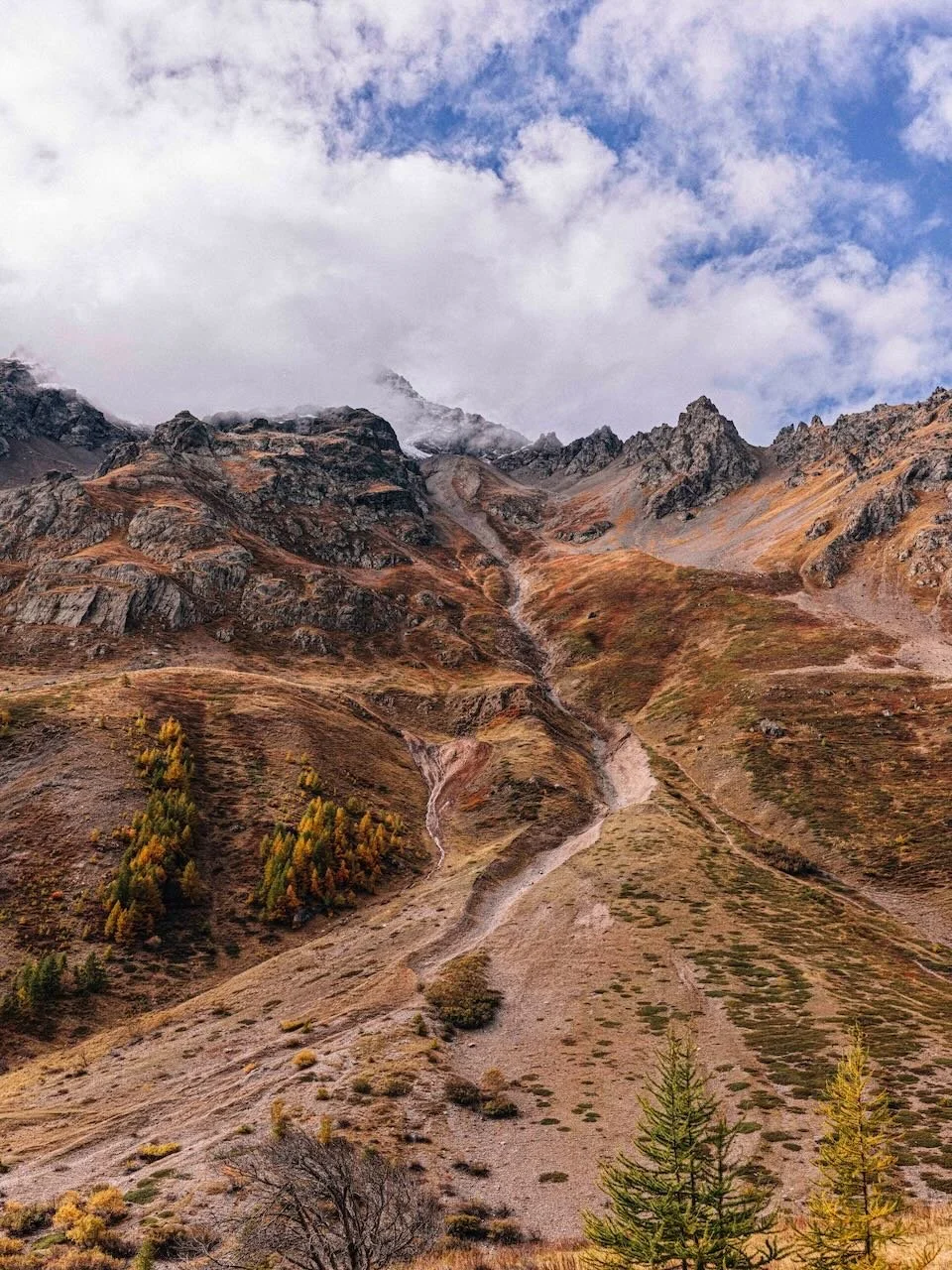 A rugged mountain landscape with steep slopes, rocky peaks, sparse vegetation, and a partly cloudy sky overhead.