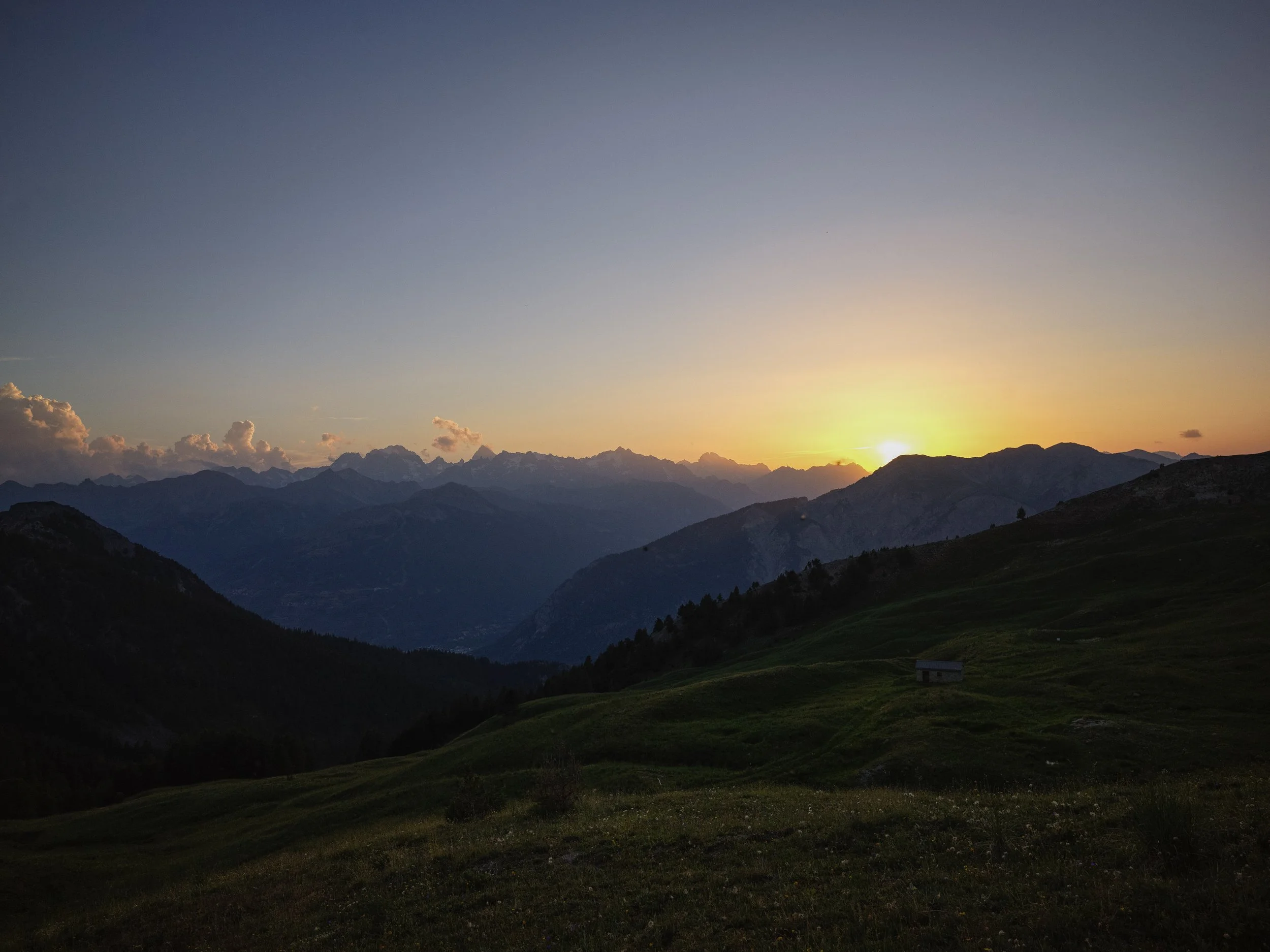 Scenic mountain landscape at sunset with green rolling hills in the foreground and distant snow-capped mountains in the background.