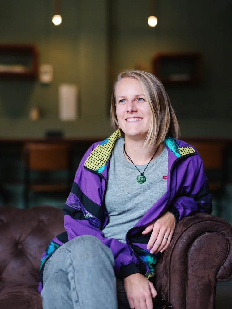 A young woman with blonde hair smiling, sitting on a brown leather couch in a dimly lit room with green walls and shelves in the background.