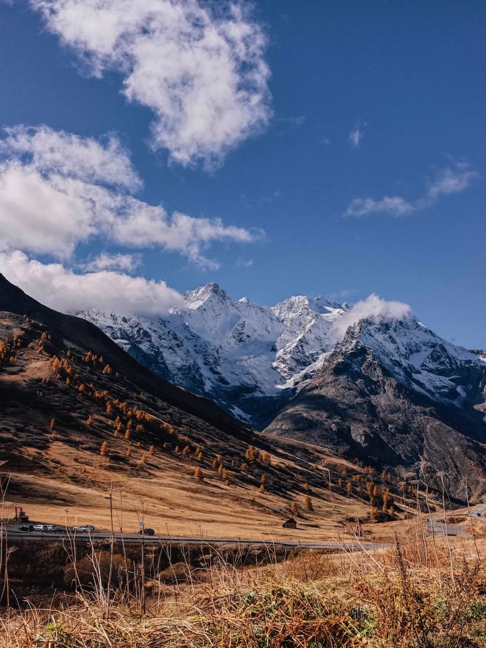 Snow-capped mountains under a blue sky with clouds, dry grass in the foreground, and a winding road with cars.