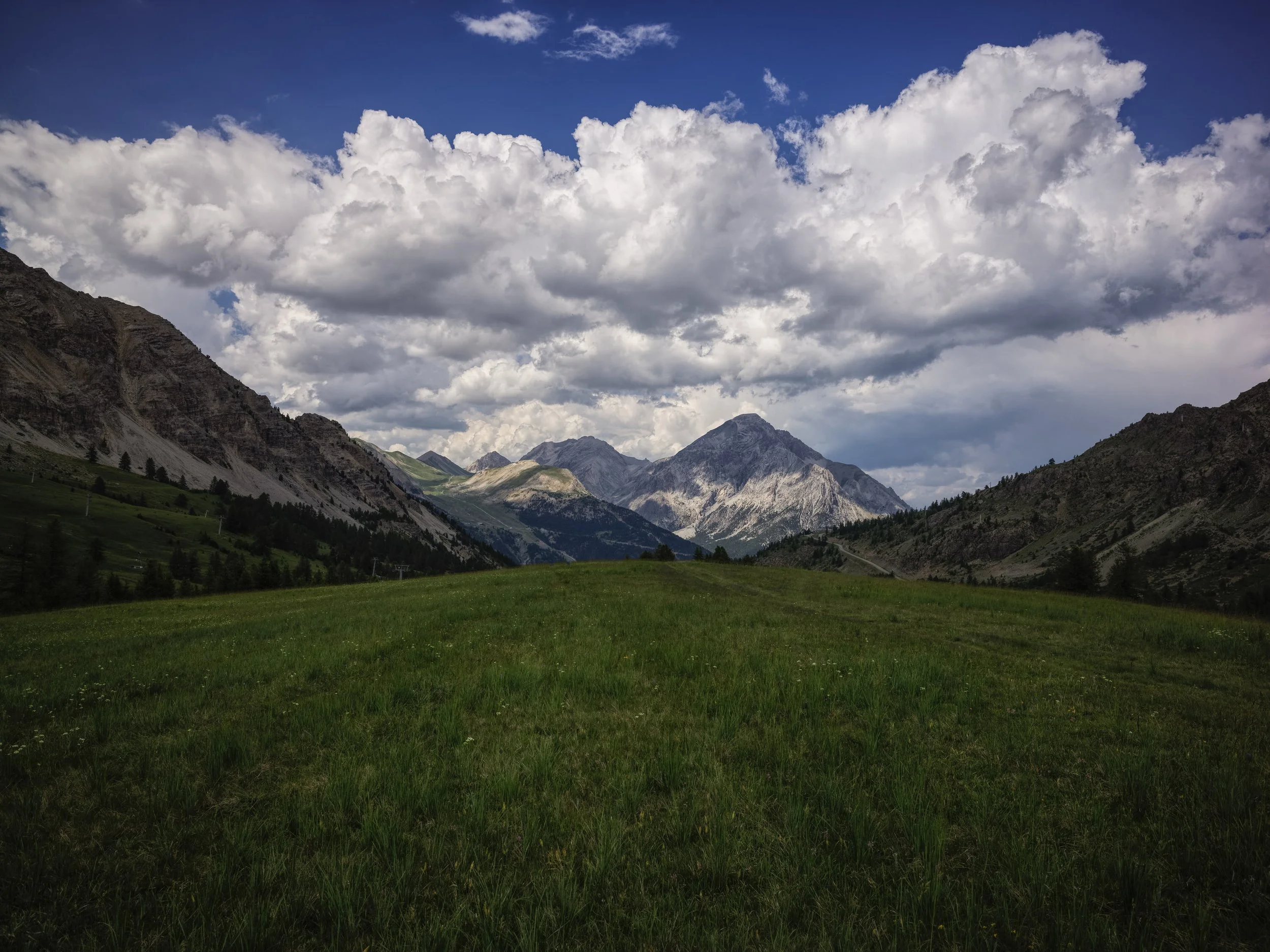 Scenic mountain landscape with lush green meadow in the foreground, rugged mountains in the midground, and dramatic cloudy sky overhead.