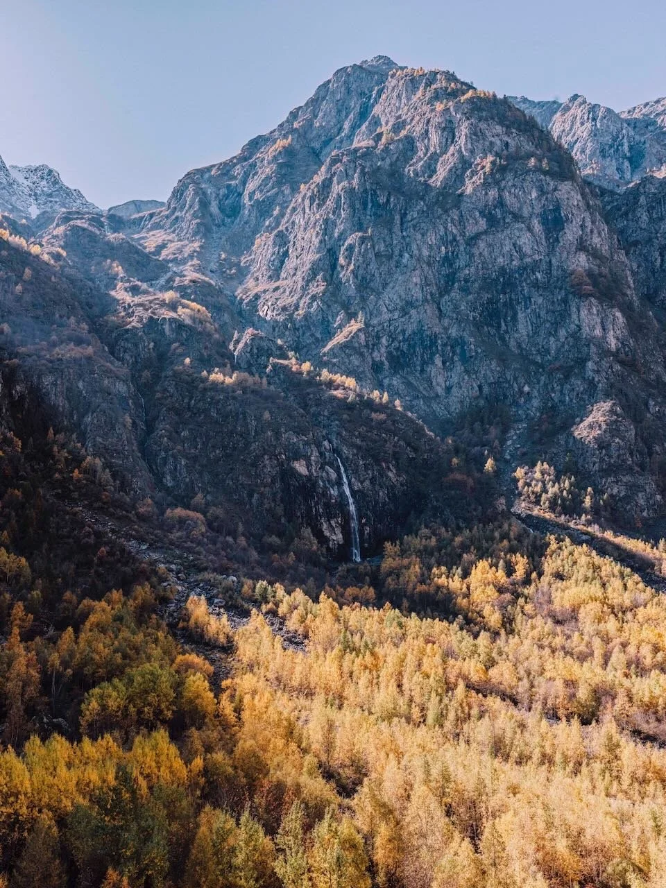 A mountain with rugged rocky slopes, snow-capped peaks, a small waterfall, and a forest of yellow-orange trees at the base.