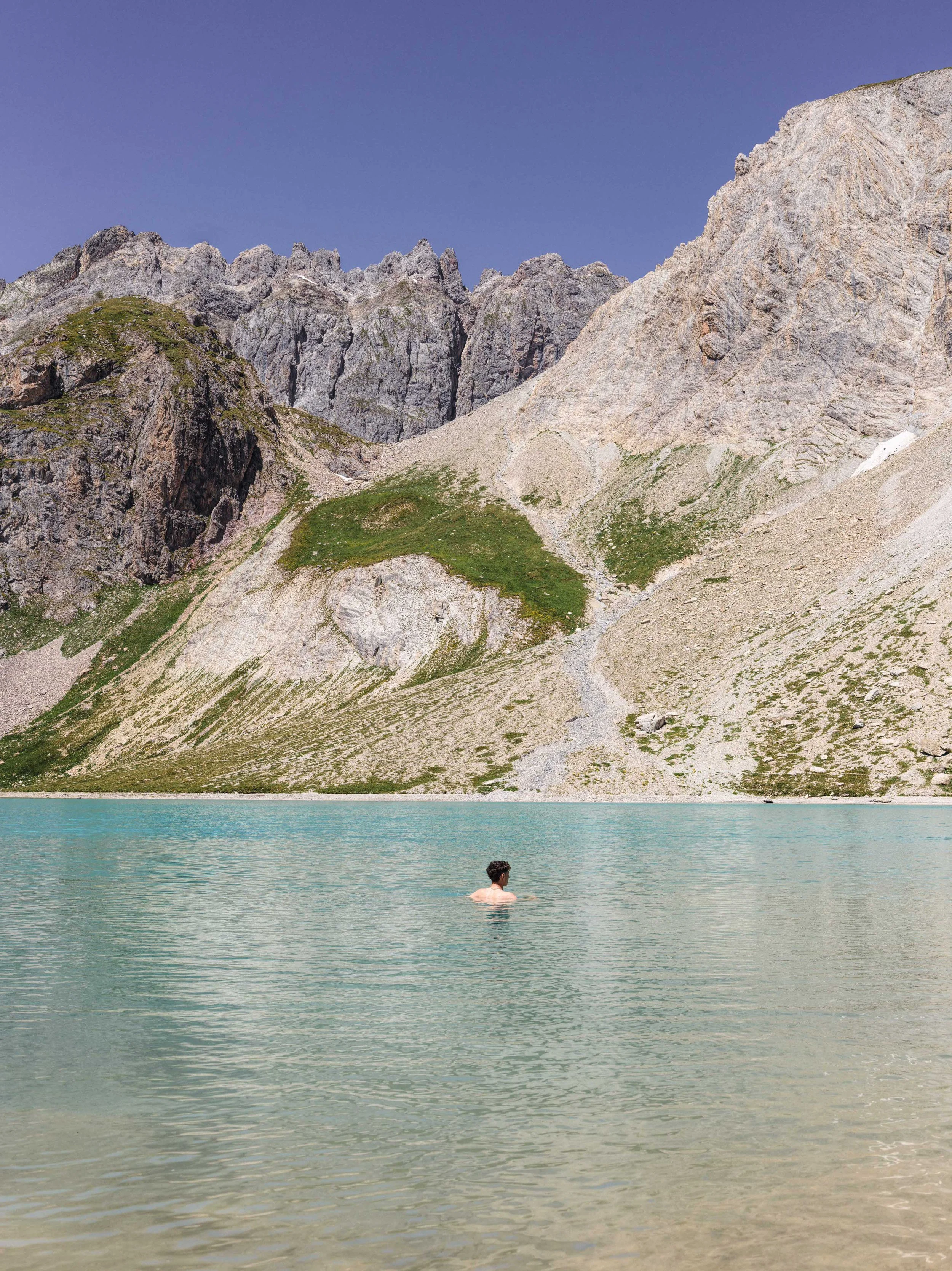 A person swimming in a turquoise mountain lake with rocky, green, and gray slopes and peaks in the background.