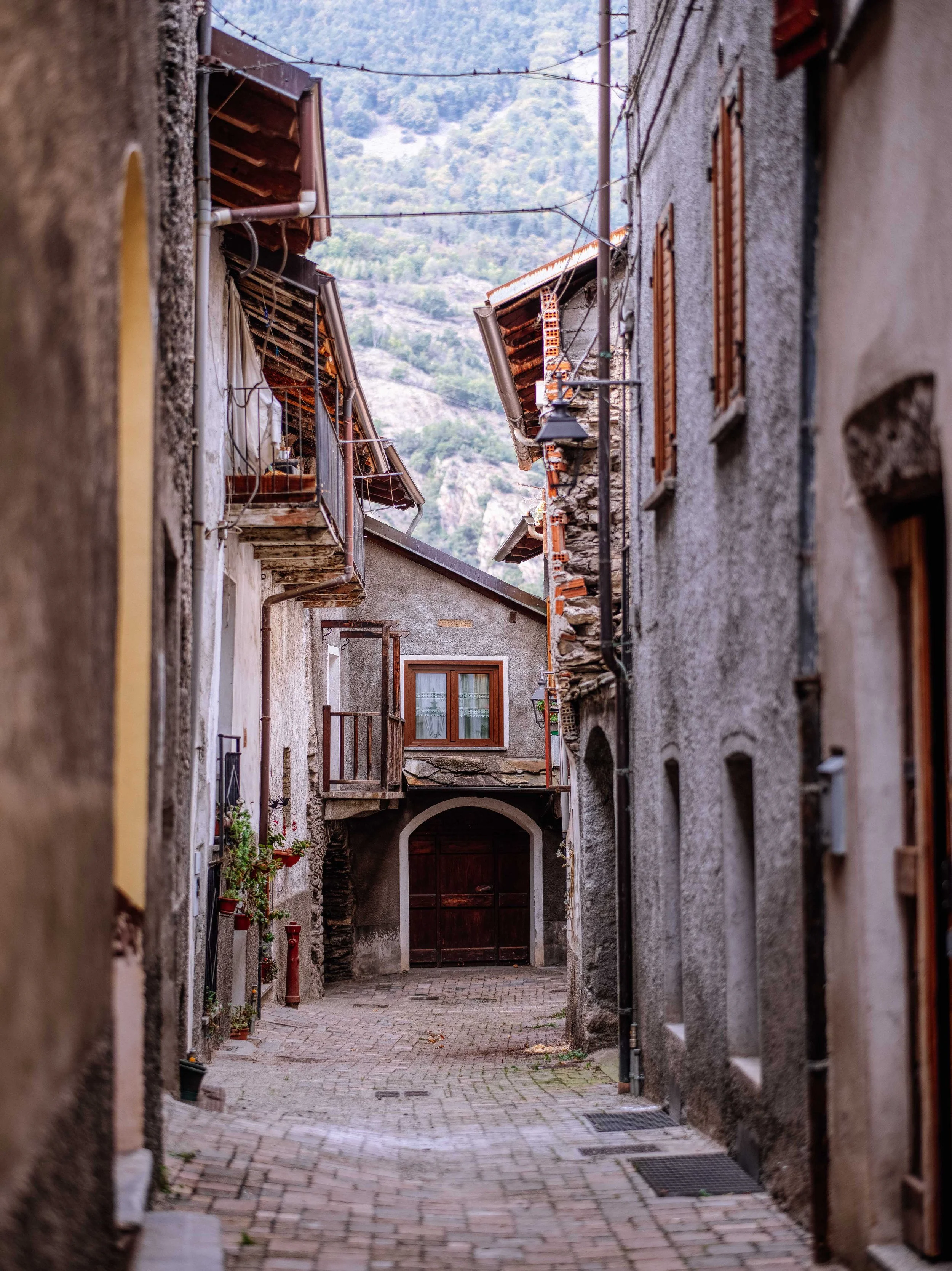 Narrow cobblestone alleyway in a mountain village with old stone and stucco buildings, wooden shutters, and hanging laundry.