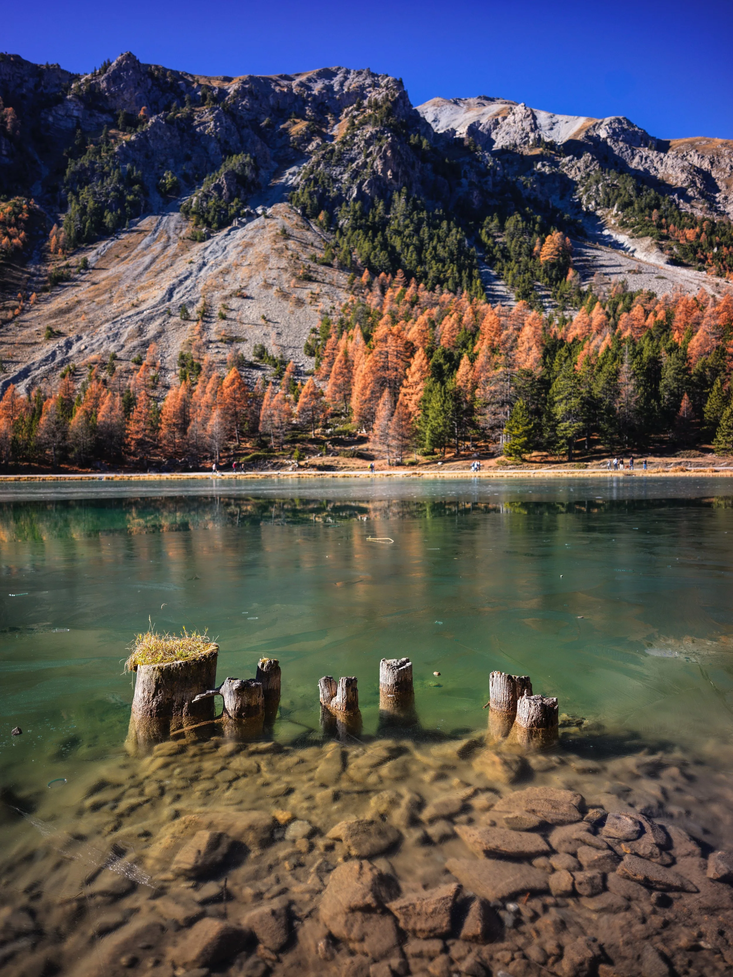 A tranquil mountain lake surrounded by autumn-colored trees and rugged, rocky mountains under a clear blue sky.