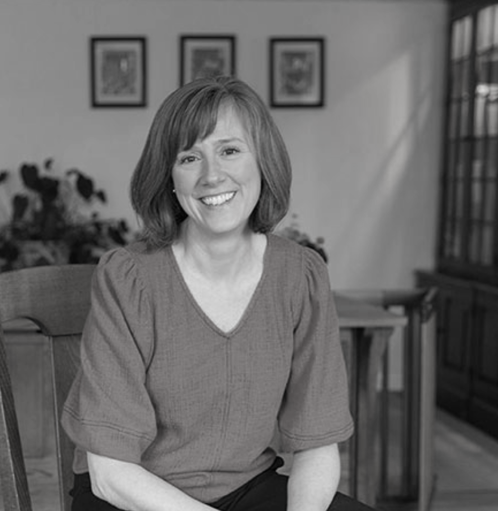 A woman with shoulder-length brown hair, smiling, wearing a red blouse, sitting on a wooden chair in a well-lit room with framed pictures on the wall and a wooden cabinet in the background. Holistic Healing, Northampton, MA.