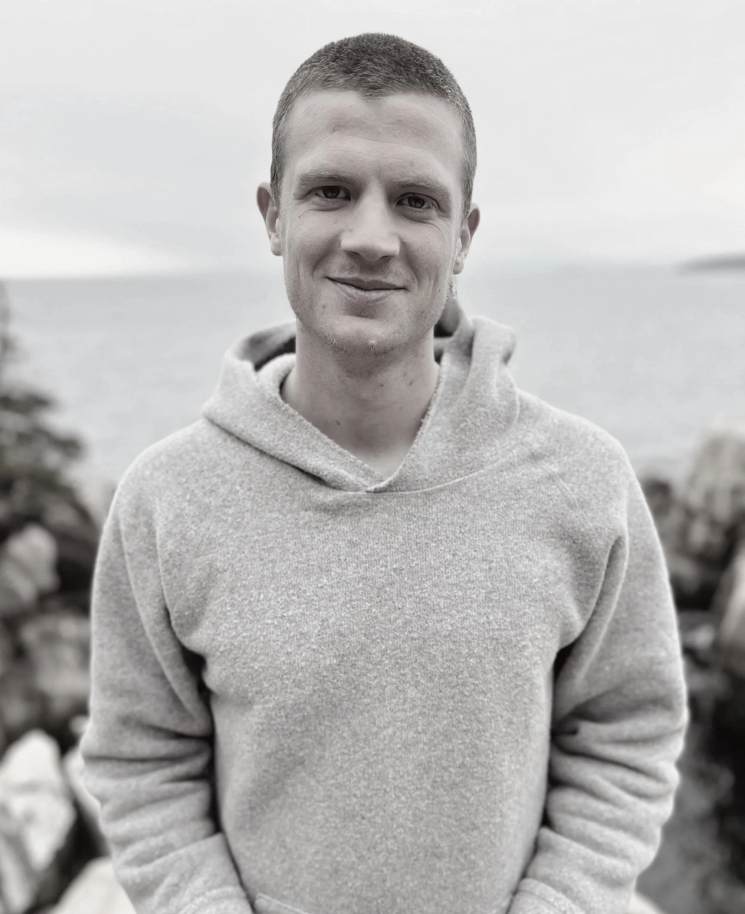 A young man, James Nepenthe, with short red hair smiling outdoors in front of a body of water and rocks, wearing a light gray hoodie. Somatic Experiencing Practitioner. 