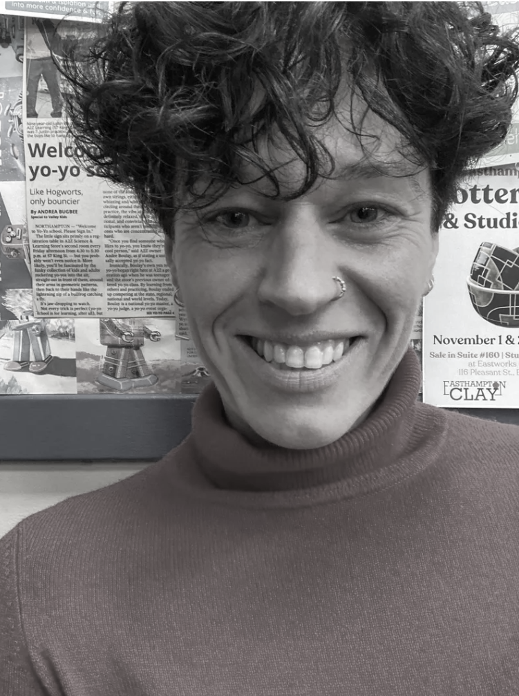 A woman with short curly dark hair, light skin, and blue eyes smiling, wearing a red turtleneck sweater and small hoop earrings with a gold nose ring. She is standing in front of a bulletin board with various posters and flyers. IFS Therapist. 