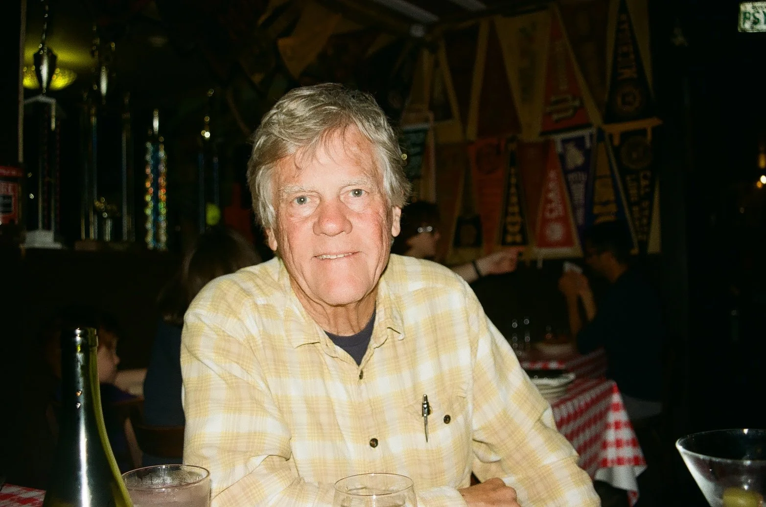 A senior man with light-colored hair, wearing a yellow plaid shirt, sitting at a restaurant table with a red and white checkered tablecloth, in a dimly lit restaurant decorated with racing banners and trophies. End of Life care, death doula. 