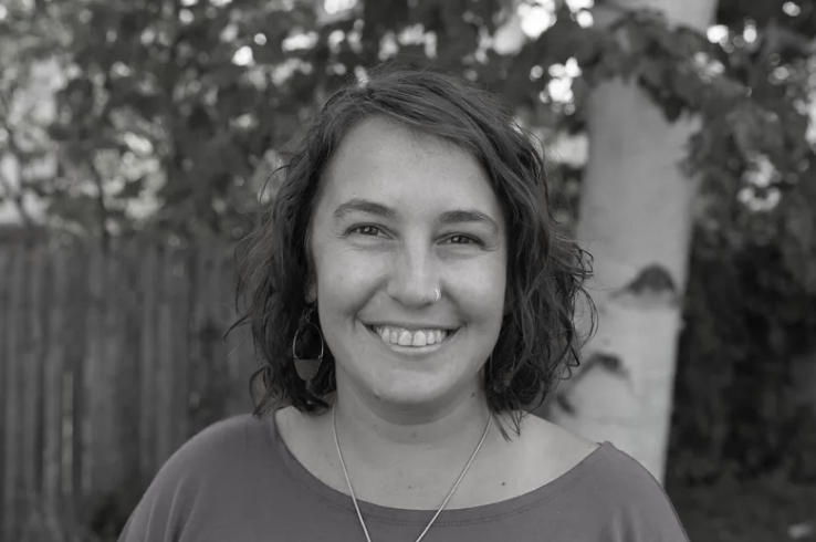 A woman with shoulder-length curly hair, wearing a purple top, standing outdoors in front of a tree and a wooden fence, smiling at the camera. IFS Therapist. 