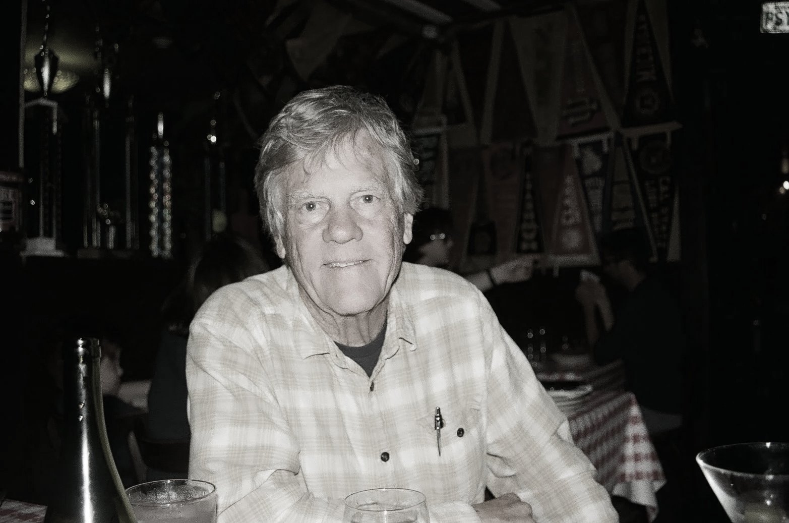 A senior man with light-colored hair, wearing a yellow plaid shirt, sitting at a restaurant table with a red and white checkered tablecloth, in a dimly lit restaurant decorated with racing banners and trophies. End of Life care, death doula. 