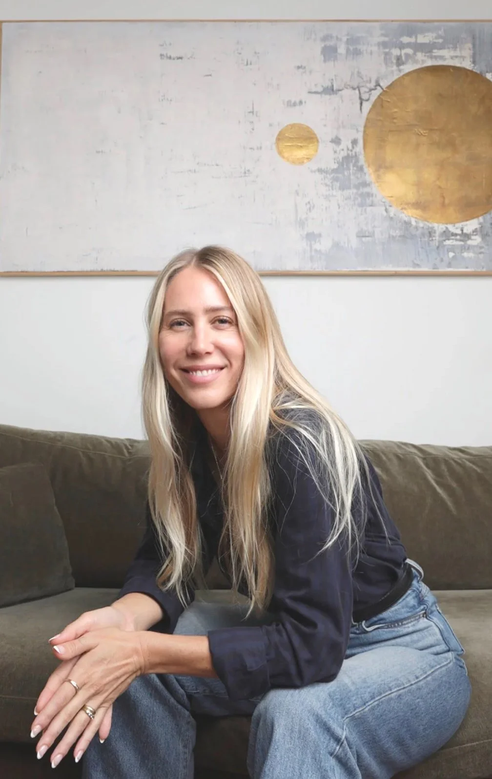 A smiling woman with long blonde hair, wearing a dark jacket and jeans, sitting on a brown couch with a white wall and abstract artwork with gold circles in the background.