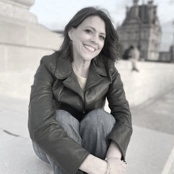 A woman, Erin Lingle, with short dark hair and red lipstick smiling, sitting outdoors on a concrete structure, wearing a green leather jacket and blue jeans. Couples Therapist.