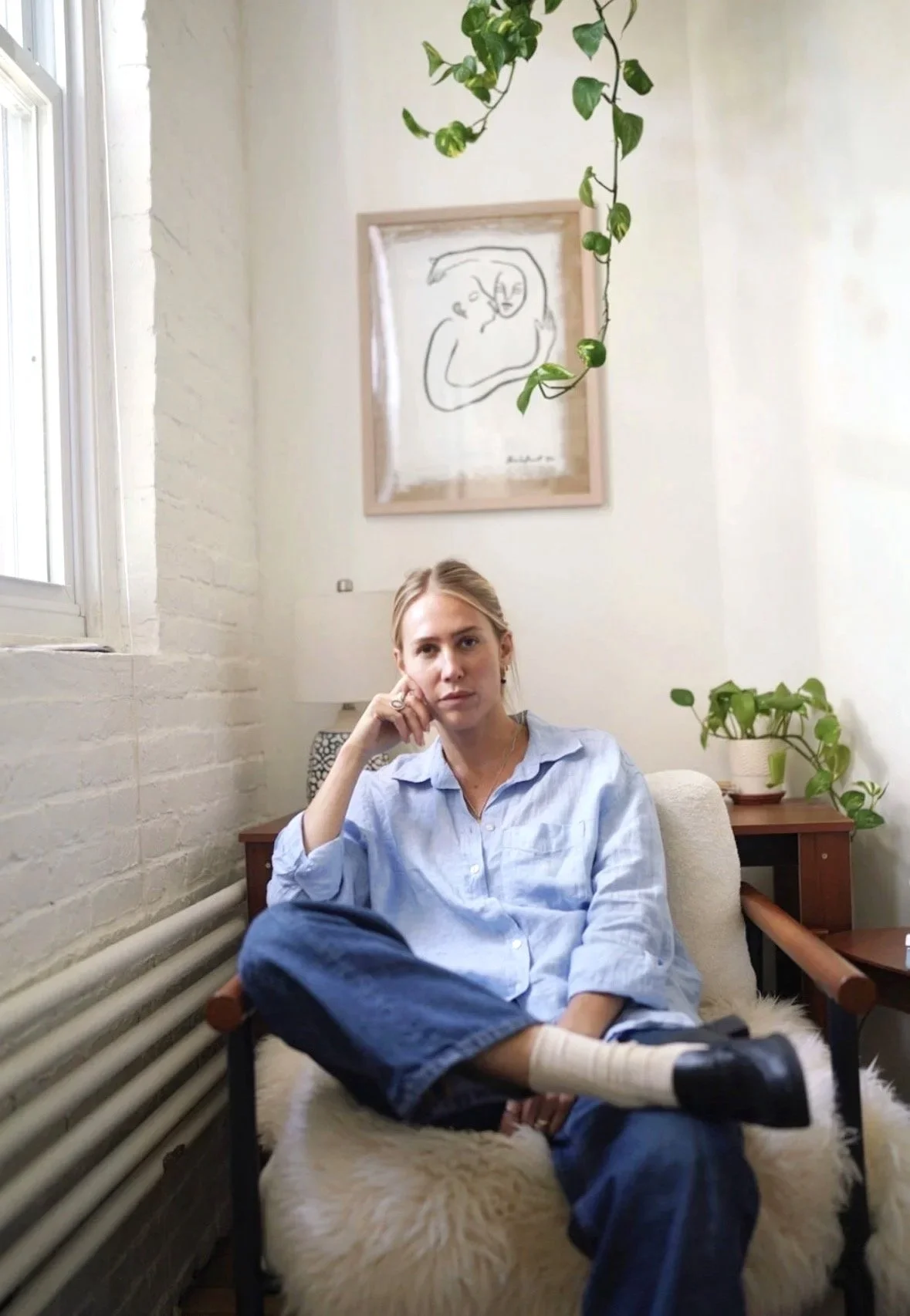 A woman sitting on a chair with her leg crossed, in a cozy room with white brick walls, a window, and houseplants. There is a framed line art drawing of a face on the wall behind her.