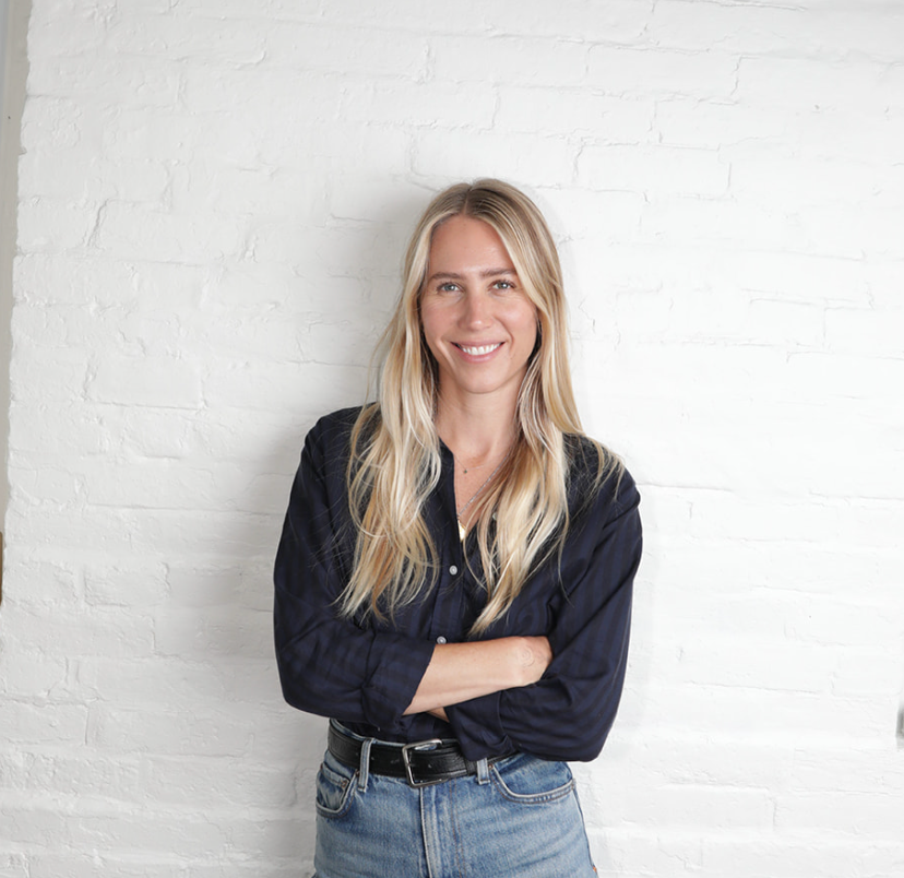 A woman with long blonde hair standing against a white brick wall, smiling, wearing a dark button-up shirt and blue jeans.