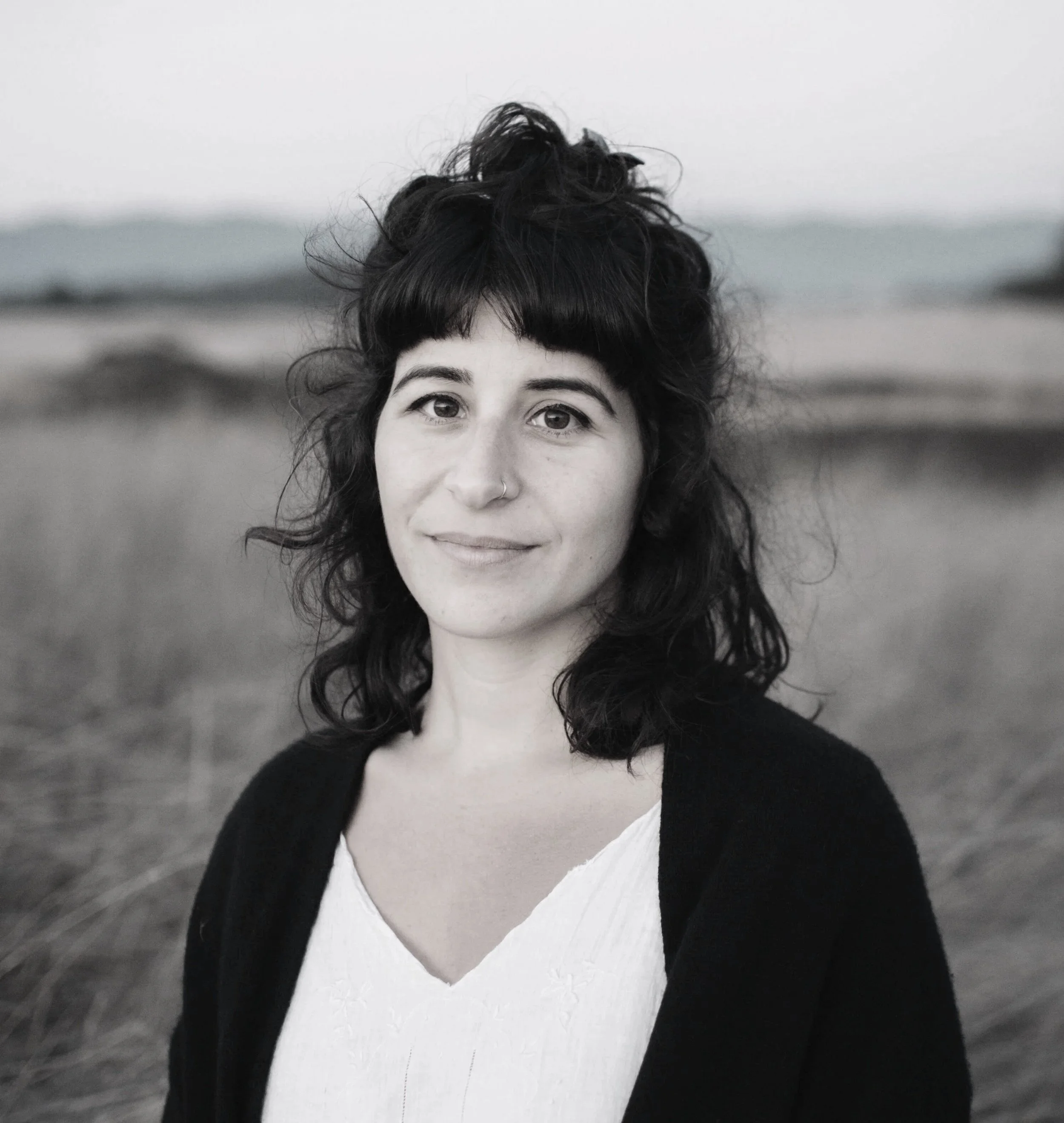 Close-up of a woman with dark, curly hair and a small nose piercing, wearing a white top and a black sweater, standing outdoors with a blurred background of sand dunes and sky.