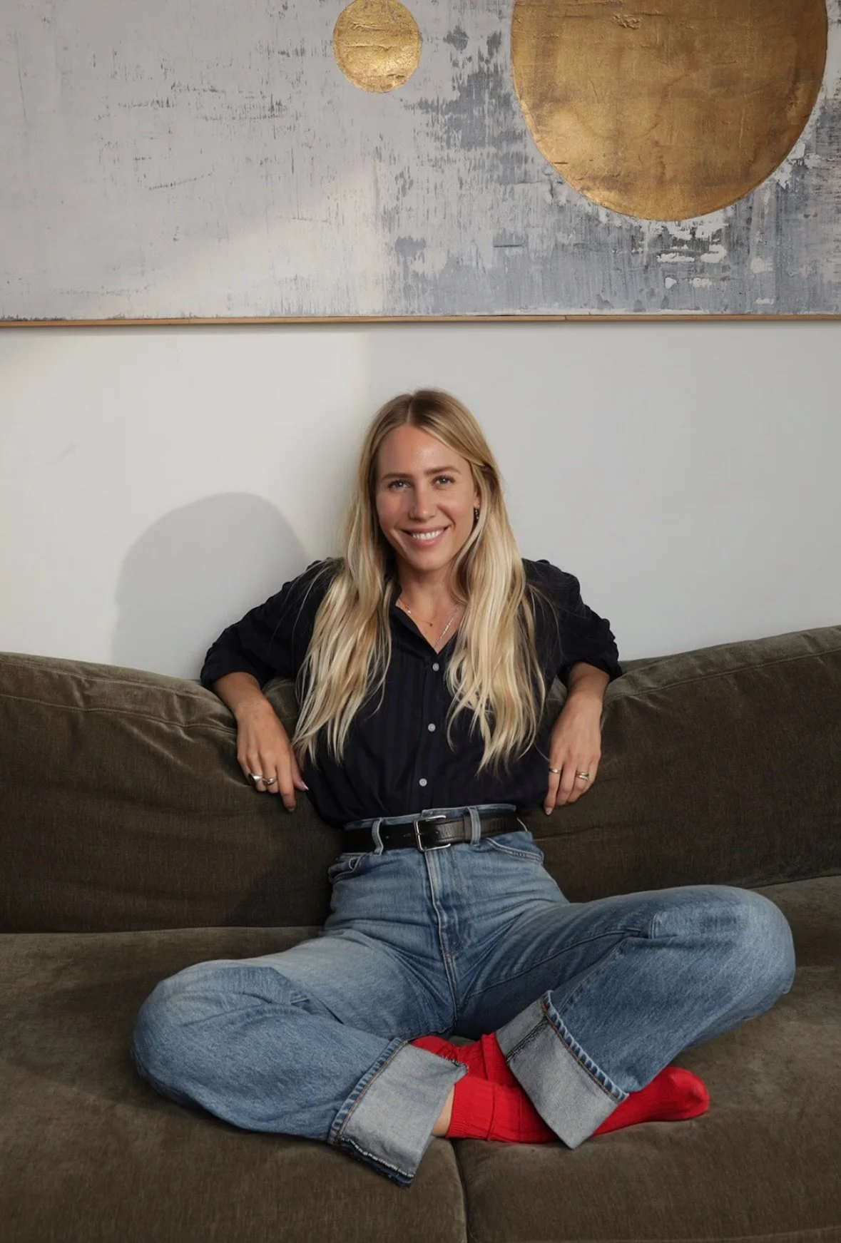 A young woman with long blonde hair sitting cross-legged on a dark brown sofa, smiling at the camera, wearing a black shirt, light blue jeans, red socks, and rings on her fingers, with modern abstract wall art featuring gold circular elements behind her.