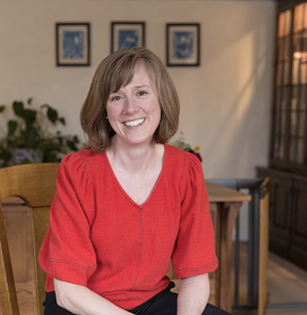 A woman with shoulder-length brown hair, smiling, wearing a red blouse, sitting on a wooden chair in a well-lit room with framed pictures on the wall and a wooden cabinet in the background. Holistic Healing, Northampton, MA.