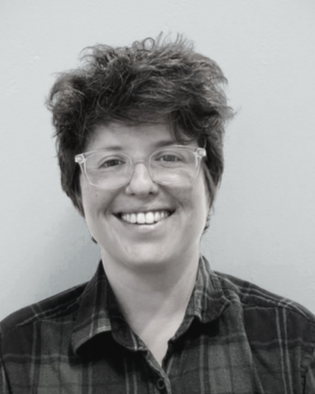 Person, JP Posnak, with short curly hair wearing glasses and a black and green plaid shirt, smiling in front of a light blue background. Couples Therapist, Northampton, MA. 