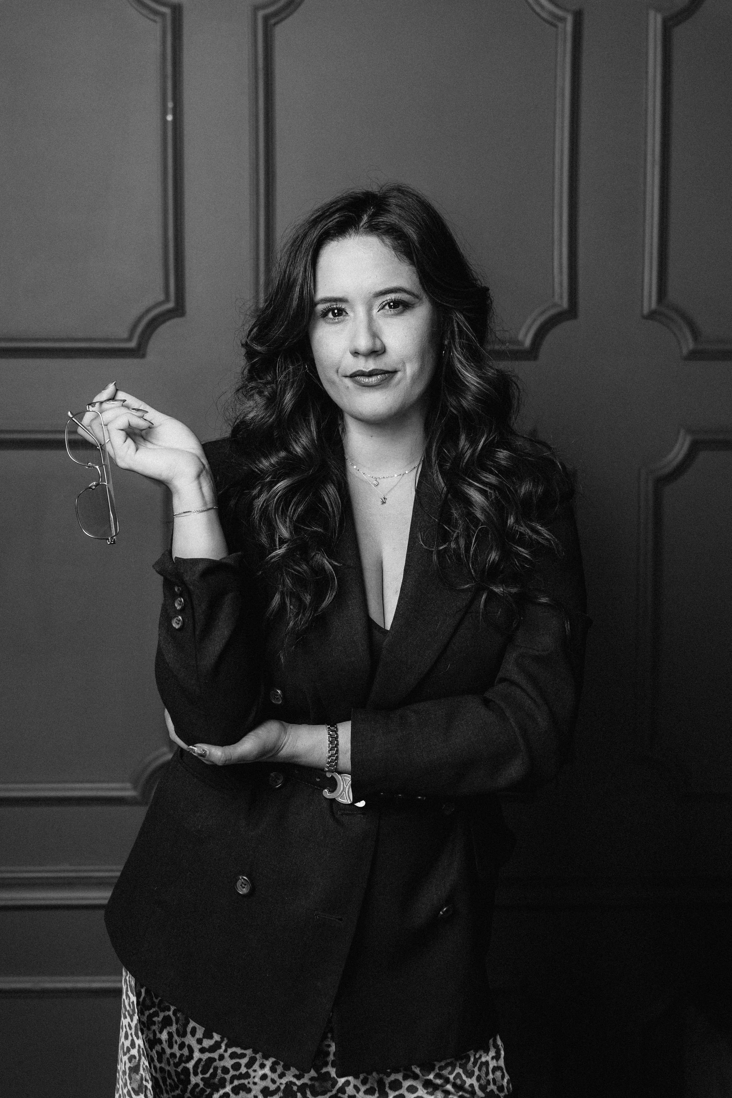 A woman with long, wavy hair holding a pair of glasses in her right hand, standing in front of a paneled wall, dressed in a dark blazer and patterned skirt, looking at the camera.