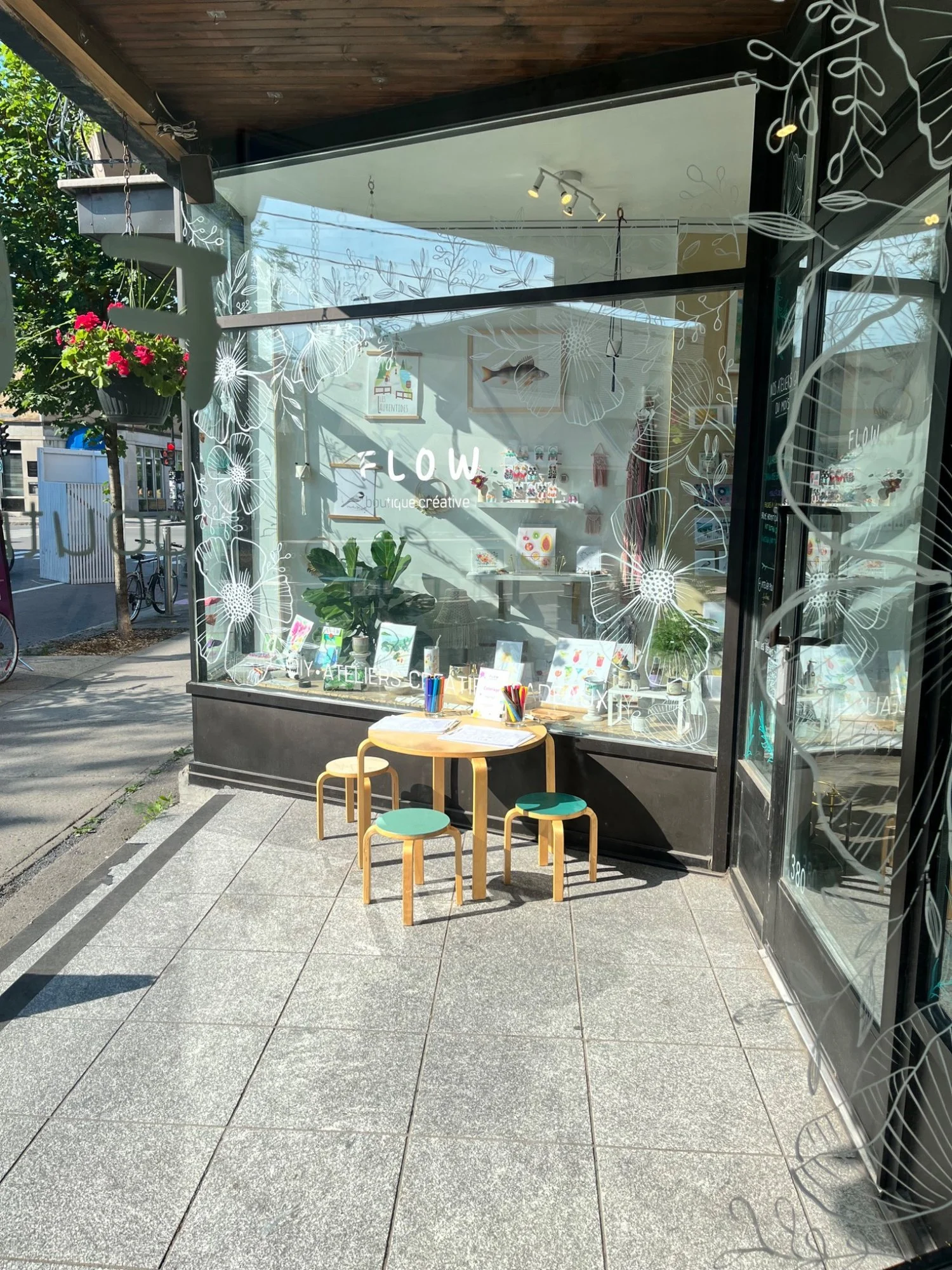 Façade d'une boutique créative avec une grande vitre décorée de dessins floraux blancs, une petite table ronde avec trois tabourets, et des plantes à l'intérieur. Dehors, une assiette en plastique avec des crayons et du papier sont posés sur la table