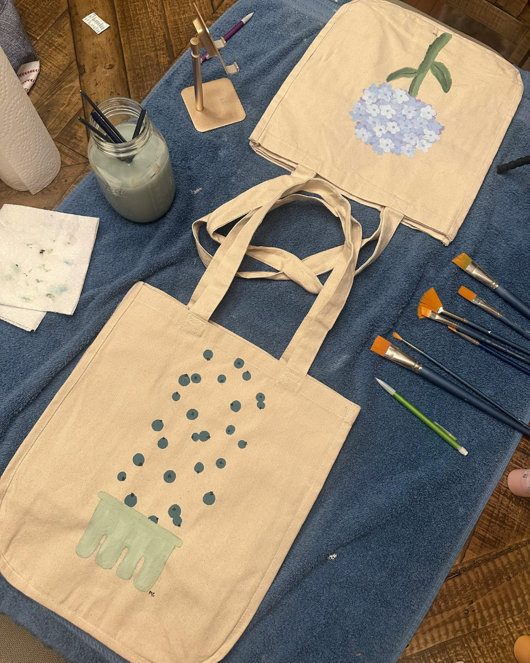 Des sacs en toile avec des motifs de fleurs, une bouteille de peinture, des pinceaux, des pinceaux à stylos, et des feuilles de papier sont disposés sur une table recouverte d'une serviette bleue.