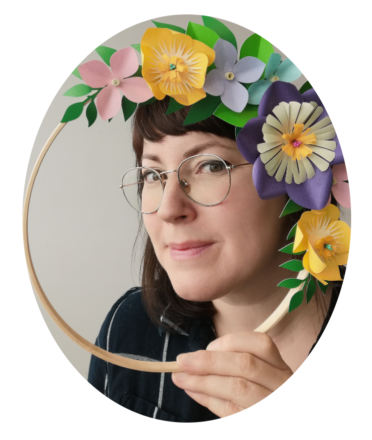 Femme portant des lunettes rondes, tenant un cadre en forme de cercle décoré de fleurs en papier de différentes couleurs, souriant légèrement.