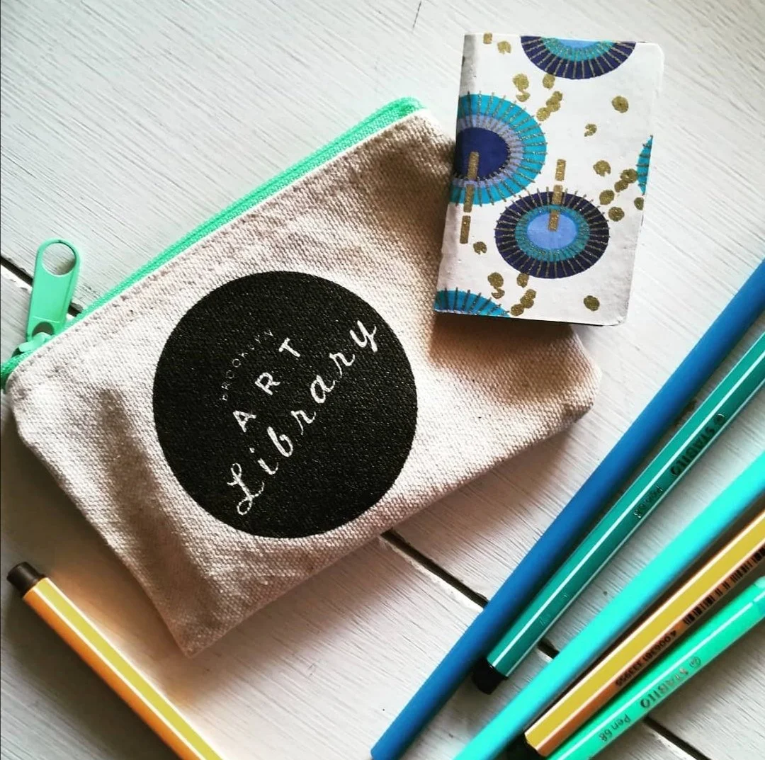 Une pochette en tissu beige avec un cercle noir portant le texte 'ART CIGARET' et divers stylos colorés à côté, sur une table en bois clair.