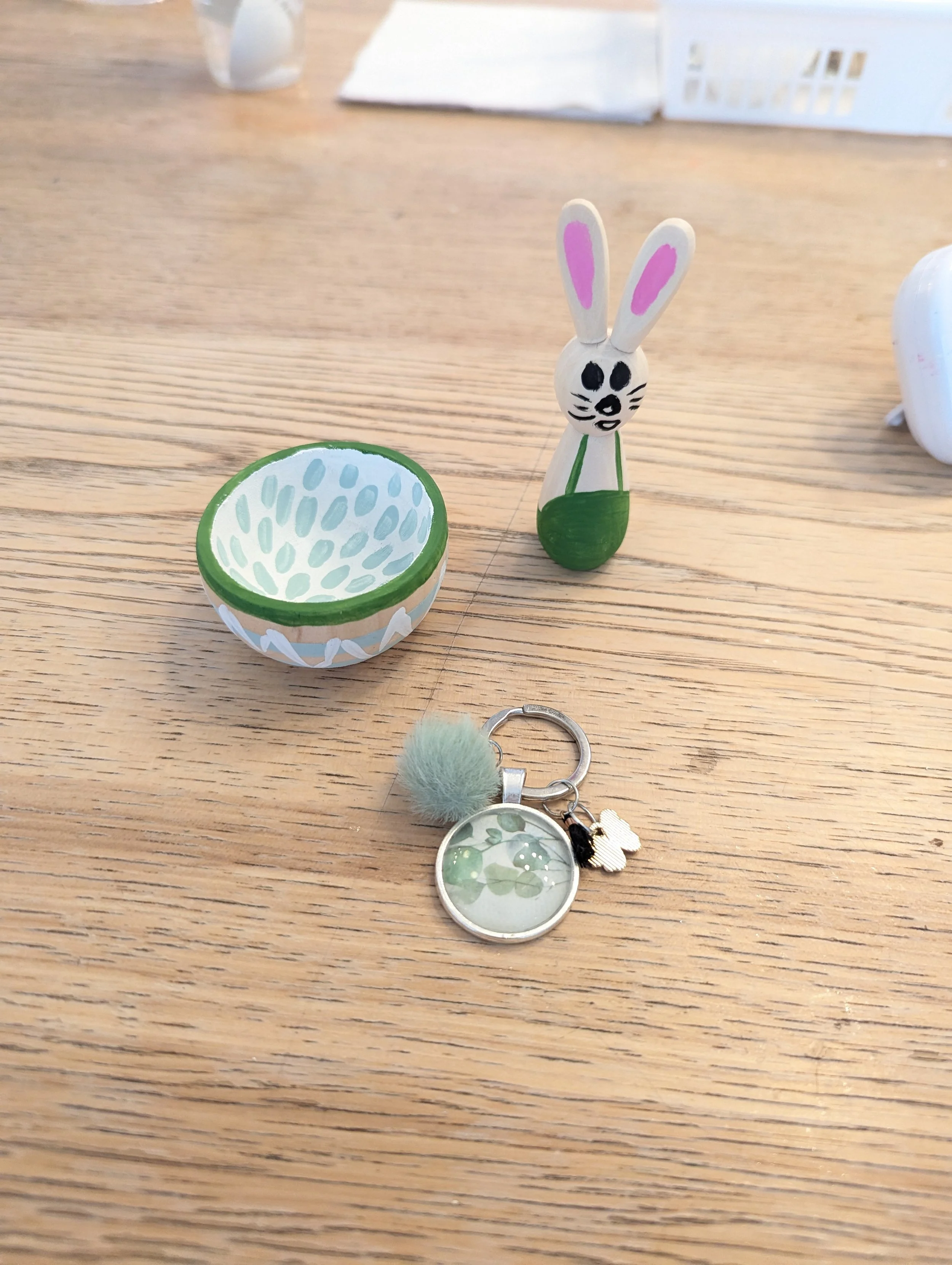 Petite figurine en bois représentant un lapin avec de longues oreilles roses, une petite coupe en bois peinte en vert et blanc, et un porte-clés avec un pompon en fourrure verte et un médaillon illustré de feuilles vertes, posés sur une surface en bo