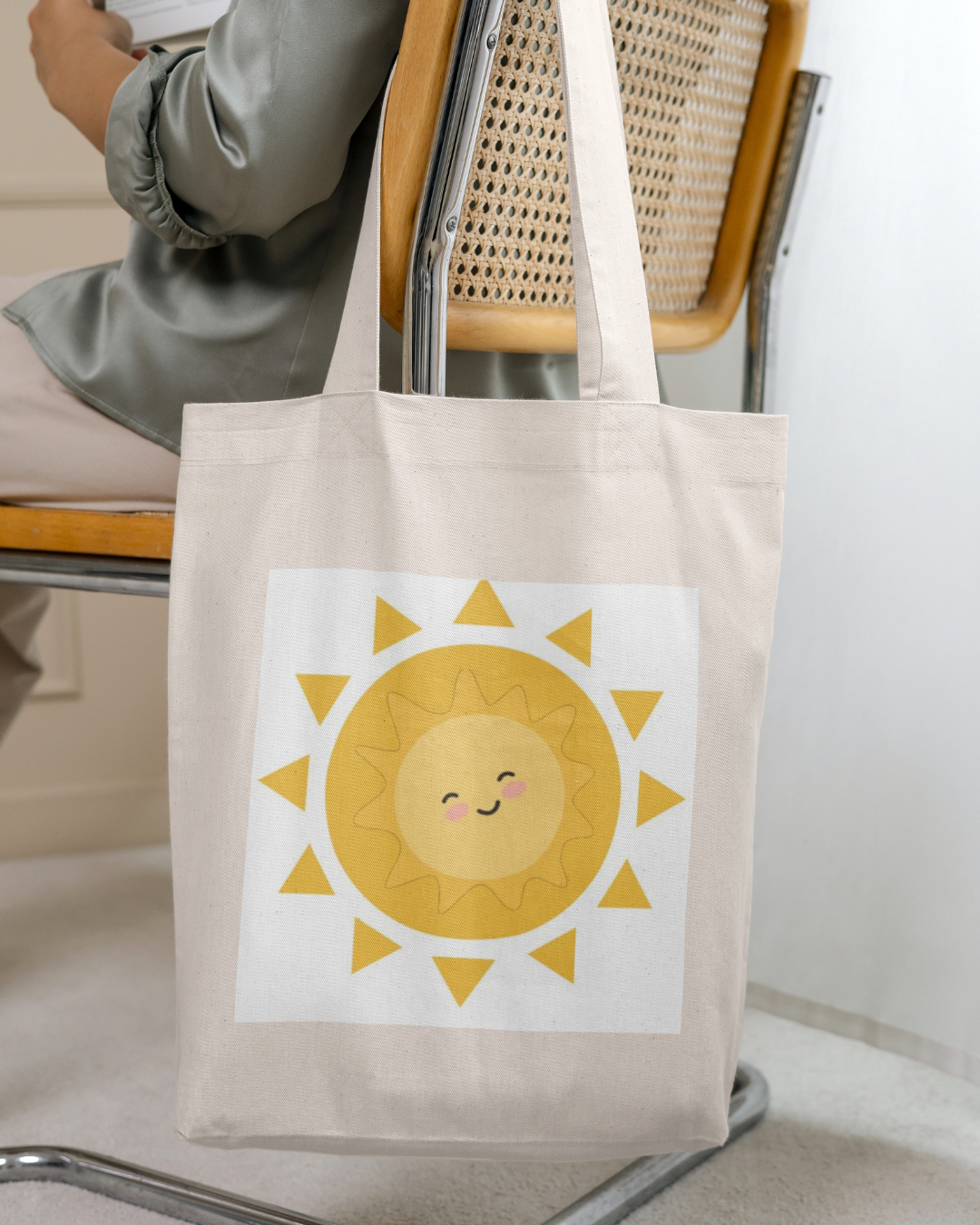 Sac en toile beige avec un dessin d'un soleil souriant coloré en jaune et orange, suspendu à la chaise en bois et métal d'une personne assise.