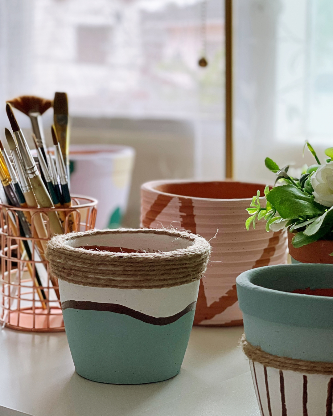 Pot de fleurs coloré, pinceaux dans un support métallique, et une plante verte avec des fleurs blanches sur une surface de table.