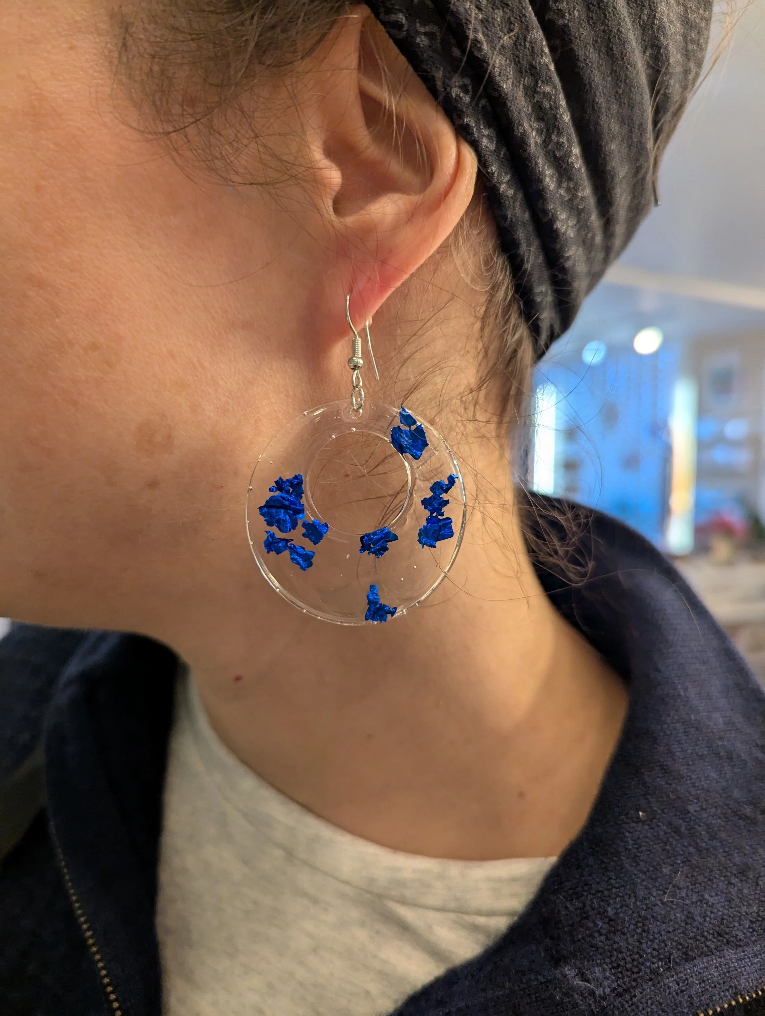 Boucle d'oreille en plastique transparent avec des morceaux de foil bleu suspendus, portée par une personne portant un bandeau noir.