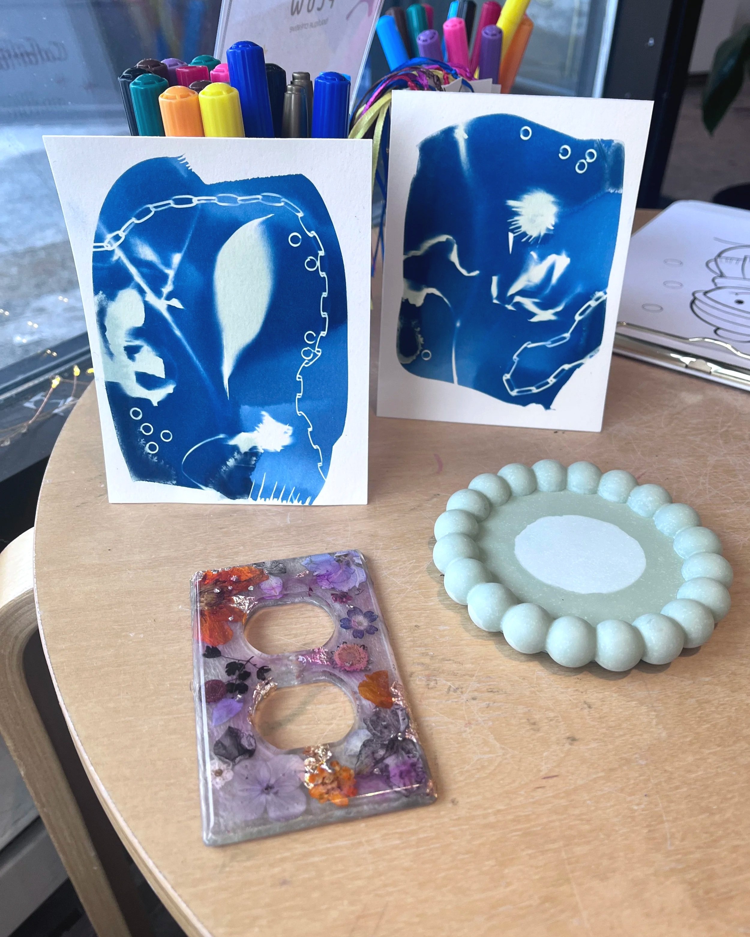 Une photo montrant des cartes de jeu avec des dessins en bleu, un bracelet en perles gris clair, un plateau avec des fleurs séchées et une boîte à bijoux, ainsi que des stylos de différentes couleurs en arrière-plan.
