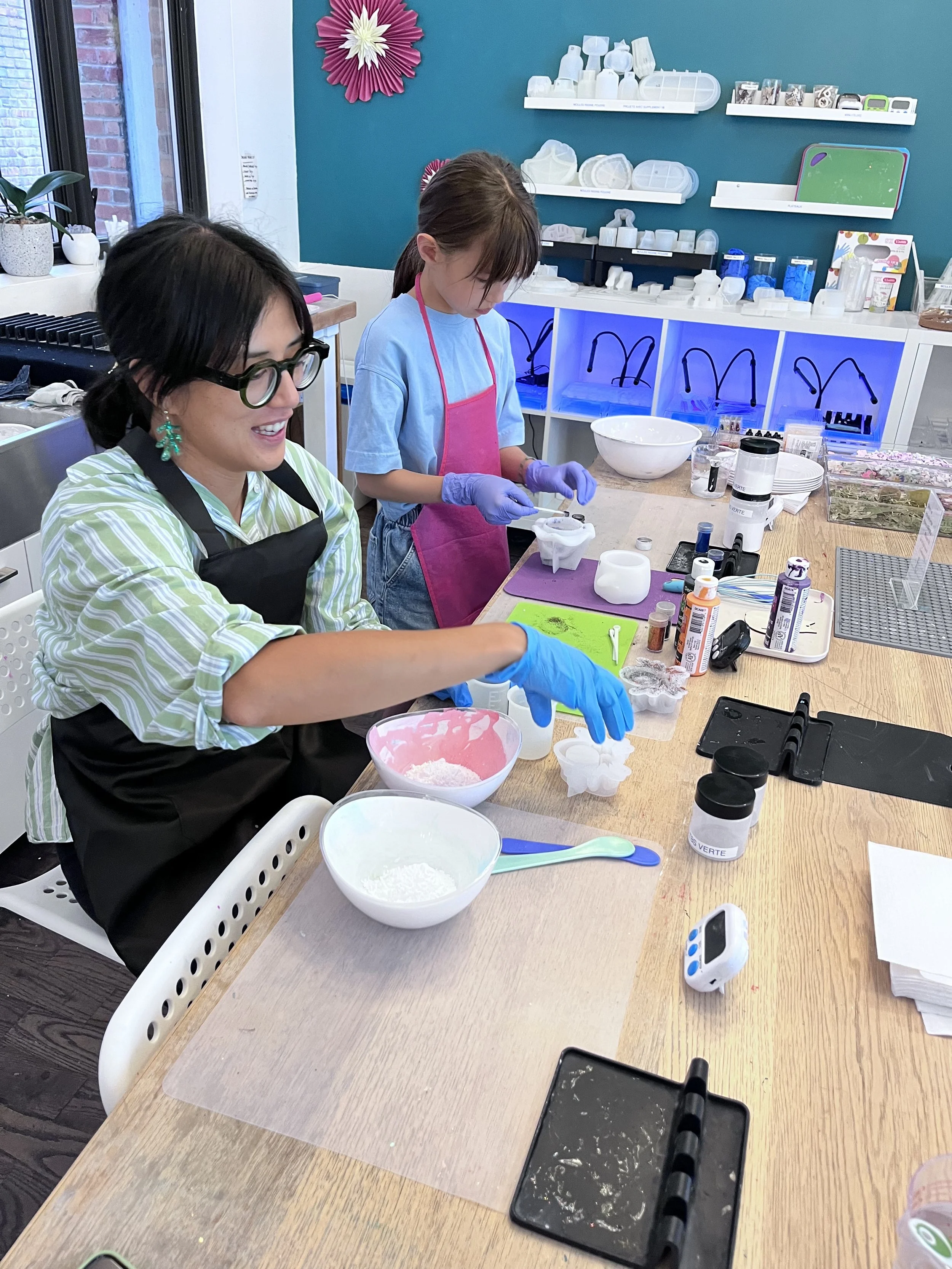 Deux personnes, une femme et une jeune fille, travaillent ensemble dans un atelier. La femme porte des lunettes épaisses, un tablier noir et une chemise à rayures vertes et blanches. La jeune fille porte un tablier rose, des gants violets et un t-shirt bleu. Elles manipulent de la pâte ou de la cire sur la table, avec divers outils, contenants et matériaux de travail autour d'elles.