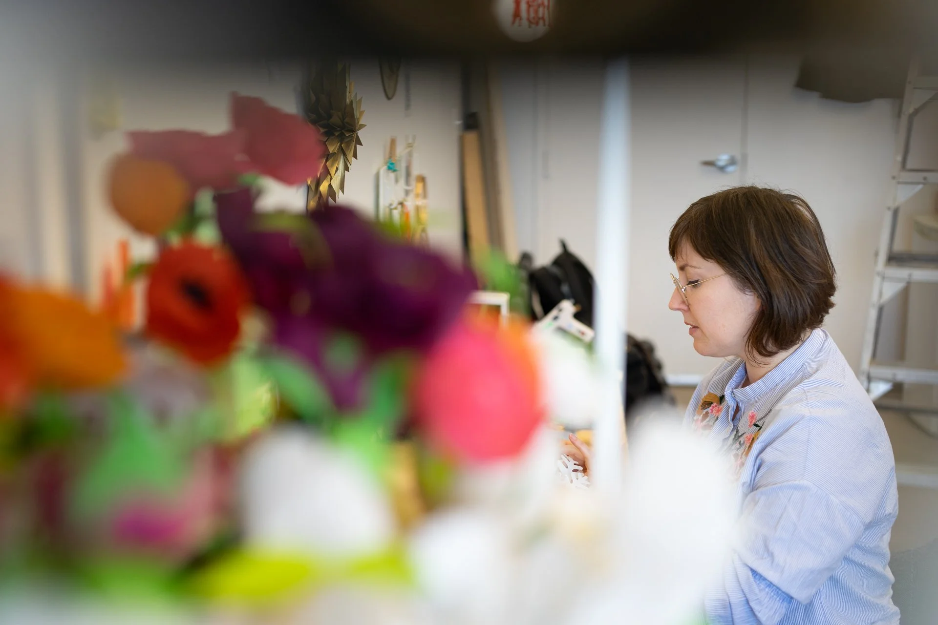 Une femme avec des lunettes et des cheveux courts bruns, concentrée, travaille dans un atelier entourée de fleurs colorées en arrière-plan.