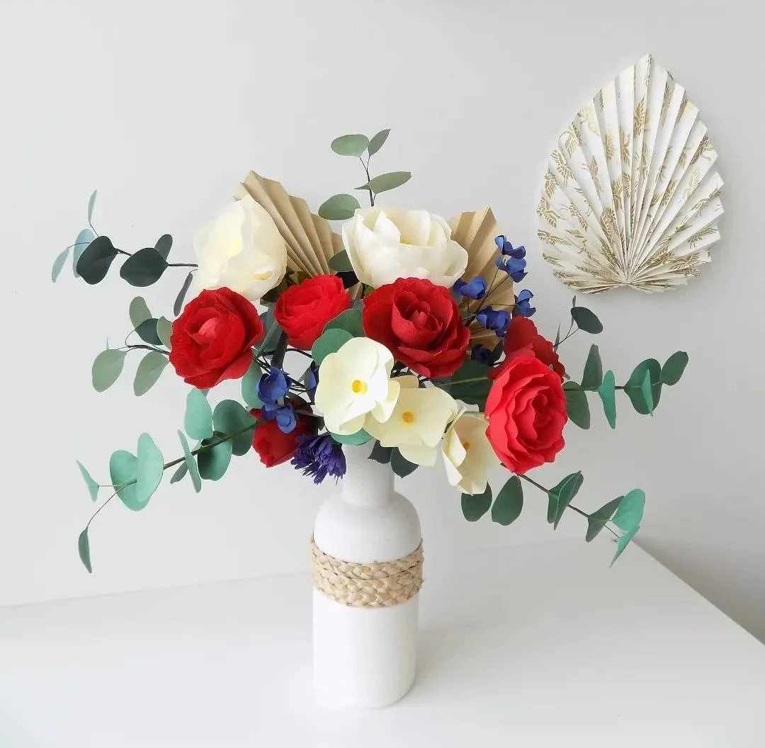 Bouquet de fleurs en papier avec des roses rouges, blanches, des hortensias bleus, dans un vase blanc avec une bande de corde, décoré avec une feuille en papier en arrière-plan.