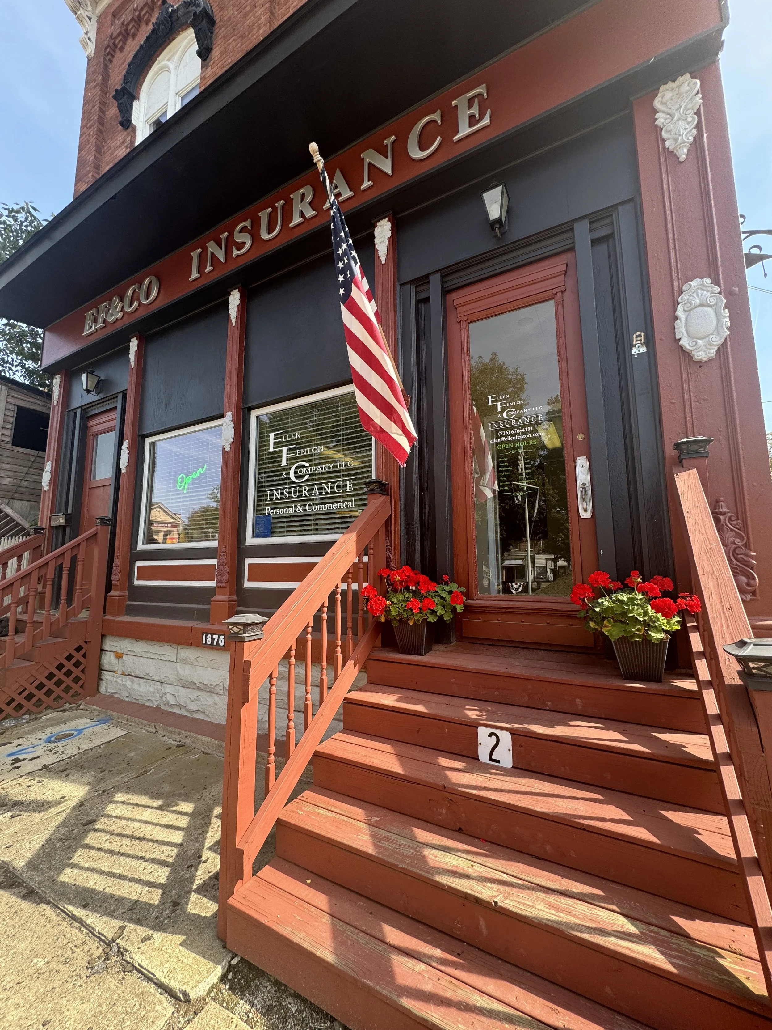 Front entrance of a red brick insurance office building with stairs, American flag, red flowers, and a neon 'Open' sign in the window