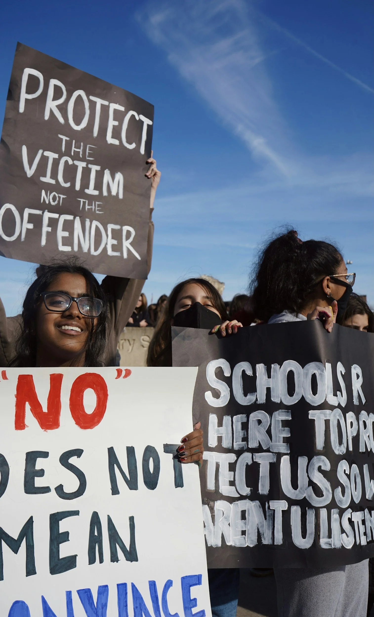 Group of people participating in a protest or rally holding signs with messages supporting victims and opposing school shootings. One sign reads "PROTECT THE VICTIM NOT THE OFFENDER" and another partially reads "SCHOOL SHOOTINGS HERE TOP".