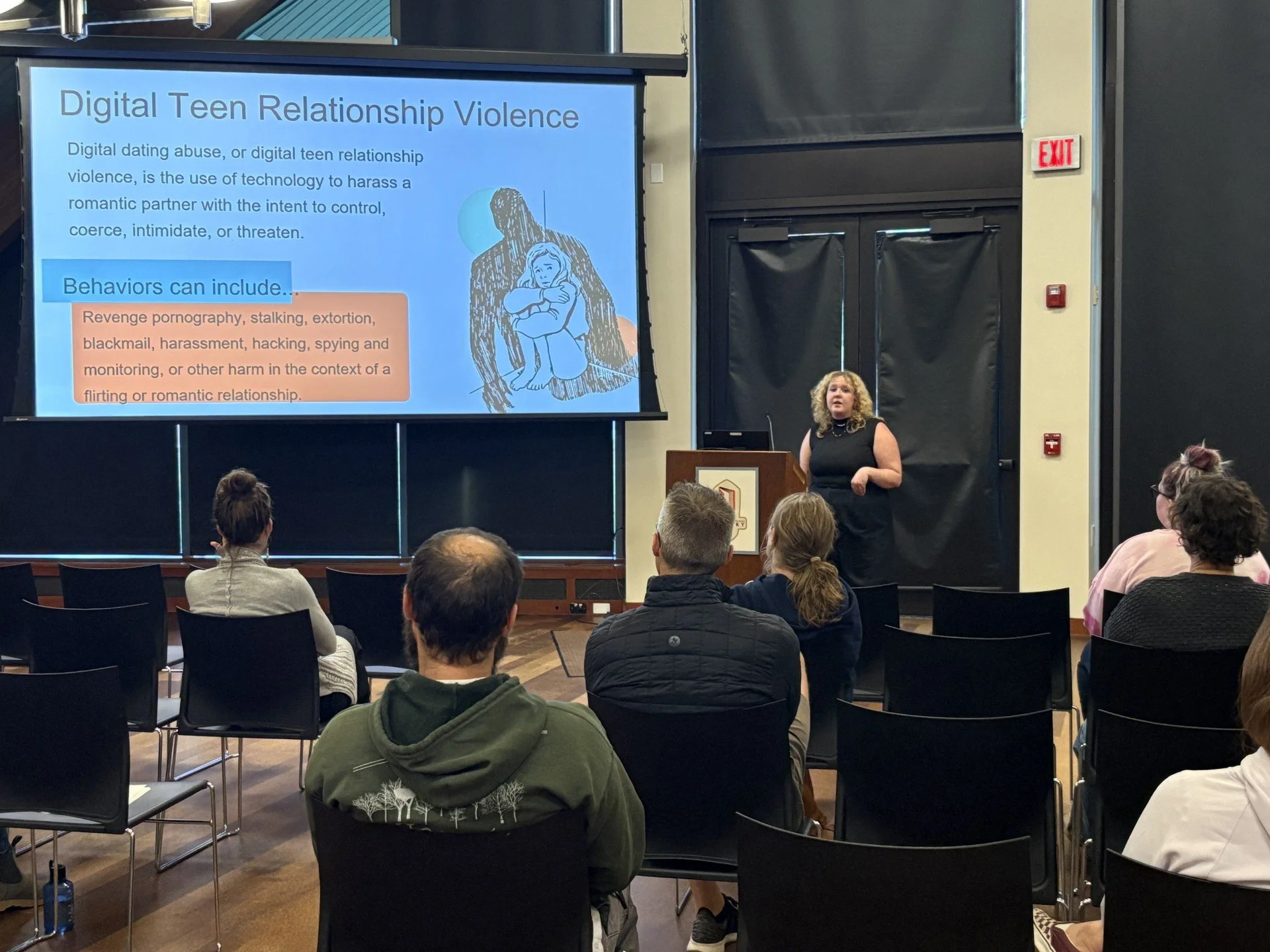 A woman giving a presentation on digital teen relationship violence to an audience seated in a room with black chairs and a large screen displaying a slide.