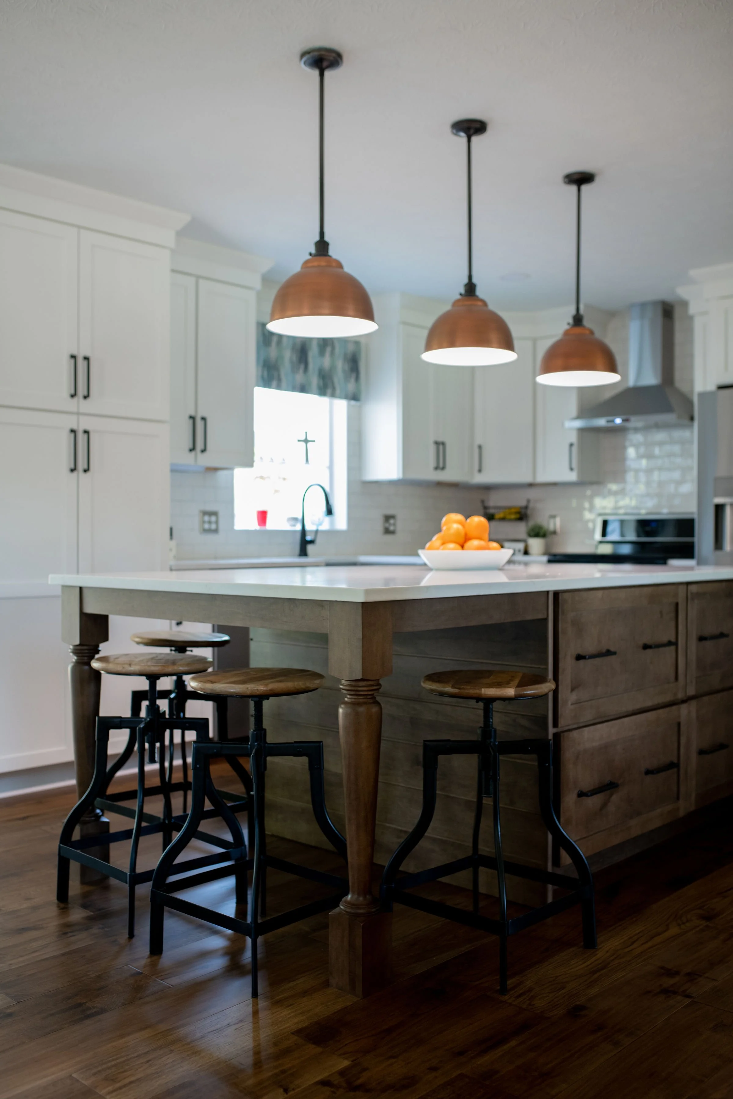 Modern kitchen with white cabinets, a large island with wooden cabinets, three copper pendant lights, and a bowl of oranges on the island. Hardwood floors and stainless steel appliances are visible.